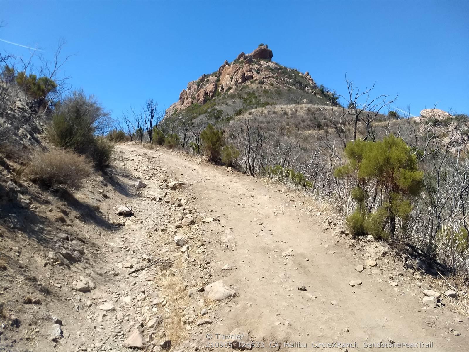Dirt path leading uphill towards a rocky mountain under clear blue sky.