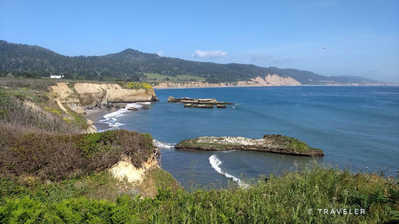 Sunny coastal view with rugged cliffs, sea stacks, and distant forested hills.