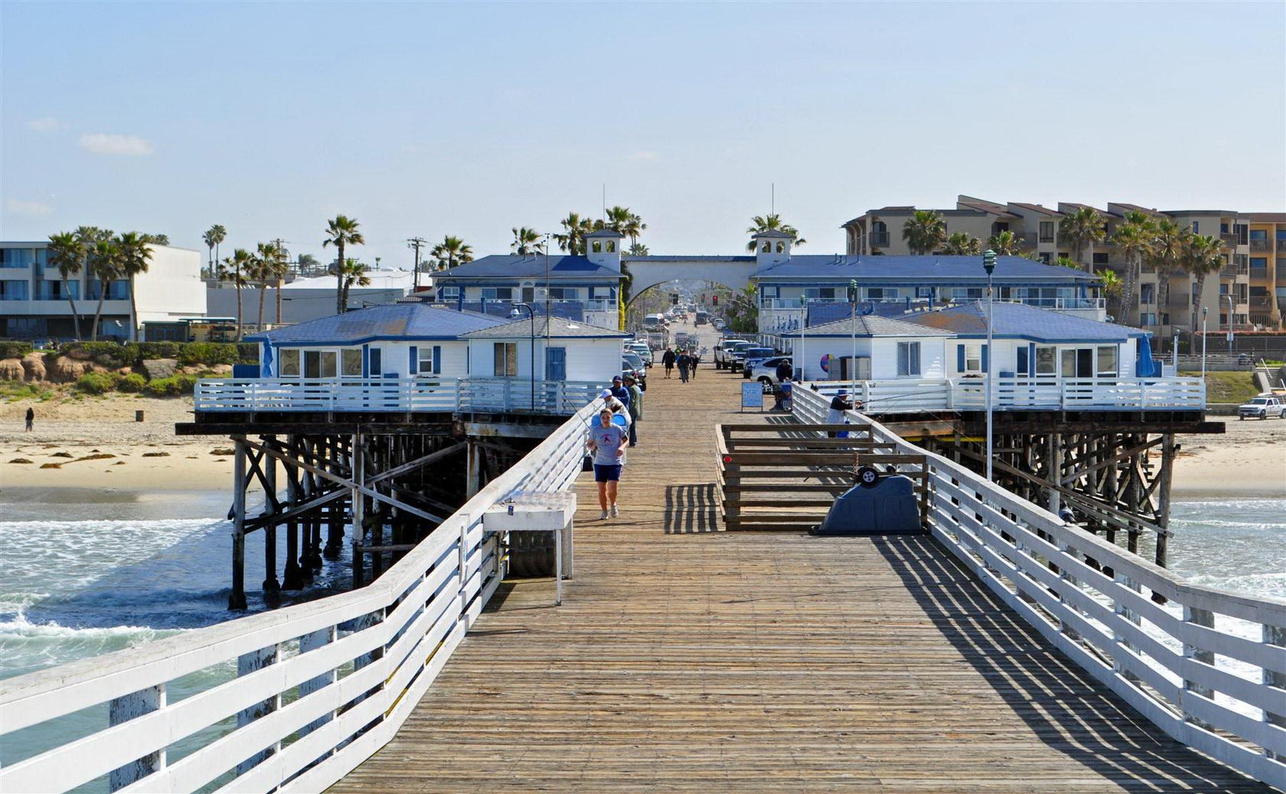 Sunny view of a wooden pier with buildings and people walking.