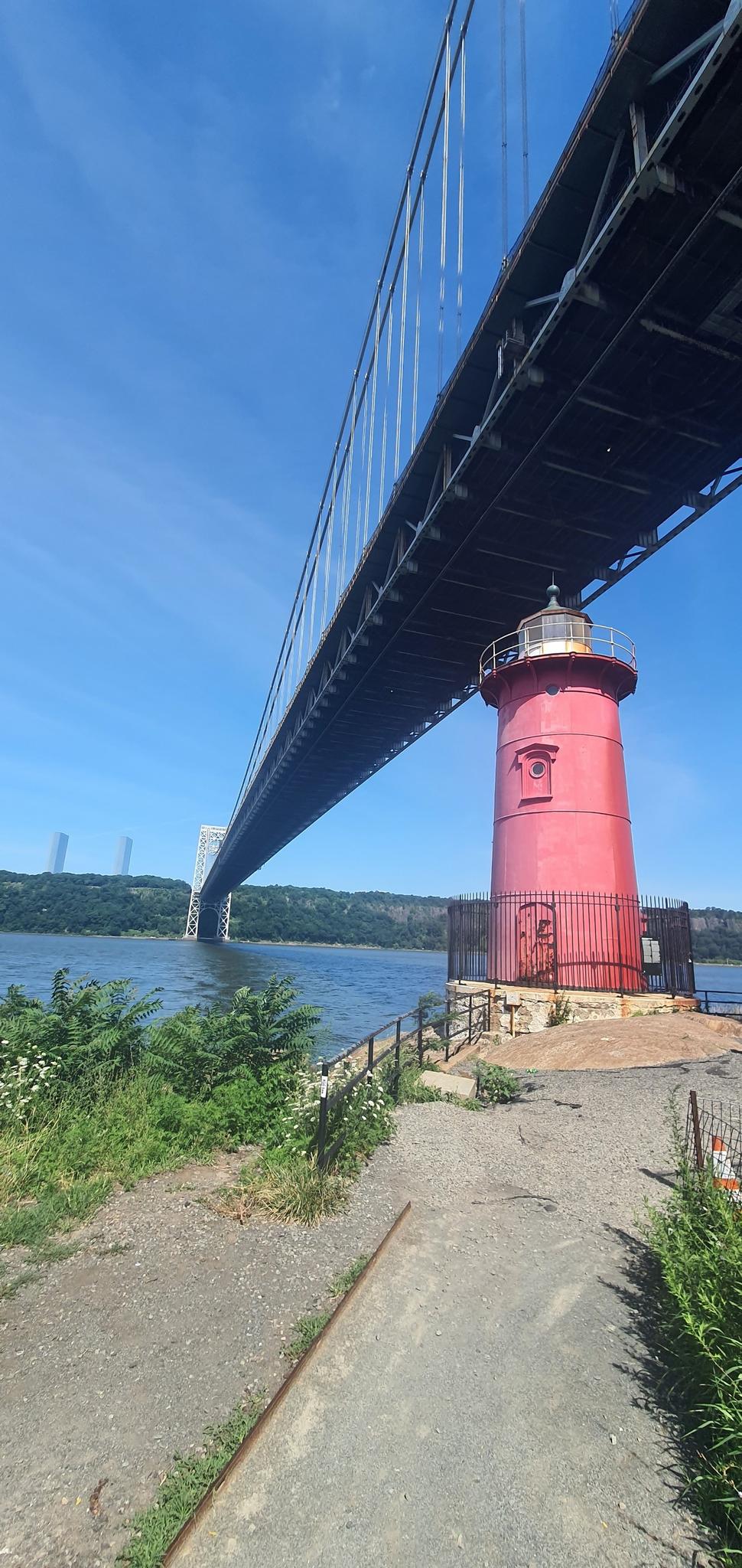 A red lighthouse stands beneath a large suspension bridge on a sunny day.