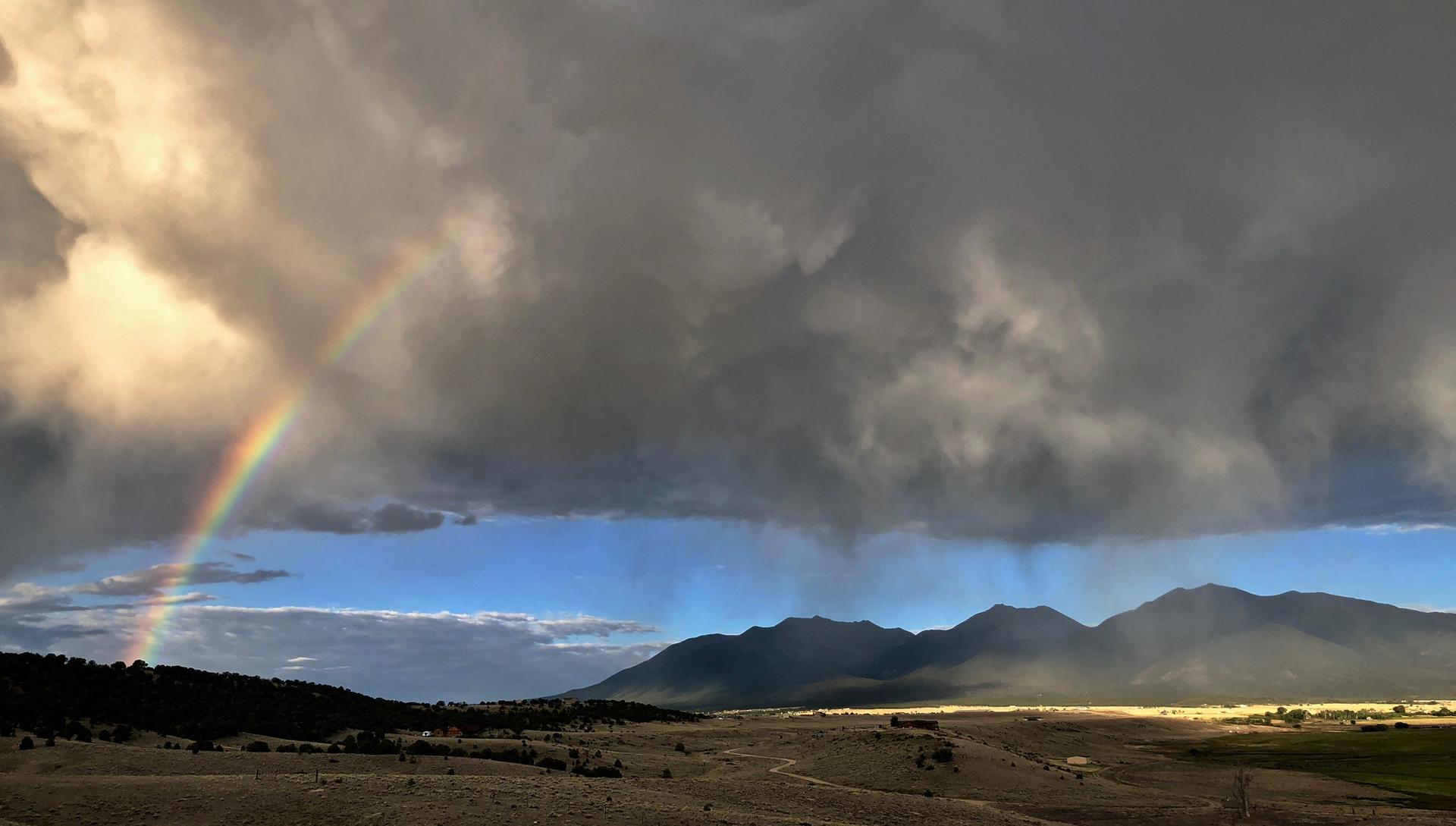A vibrant rainbow arches over a sunlit valley with dark mountains under stormy skies.