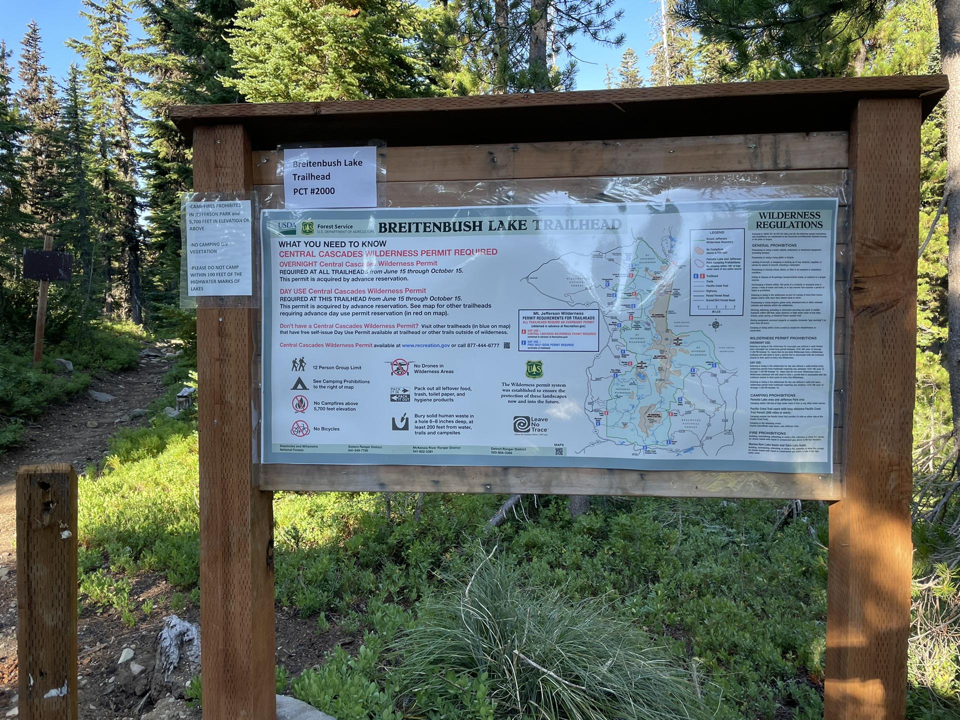 Wooden trailhead sign with map and information in a sunny forest.