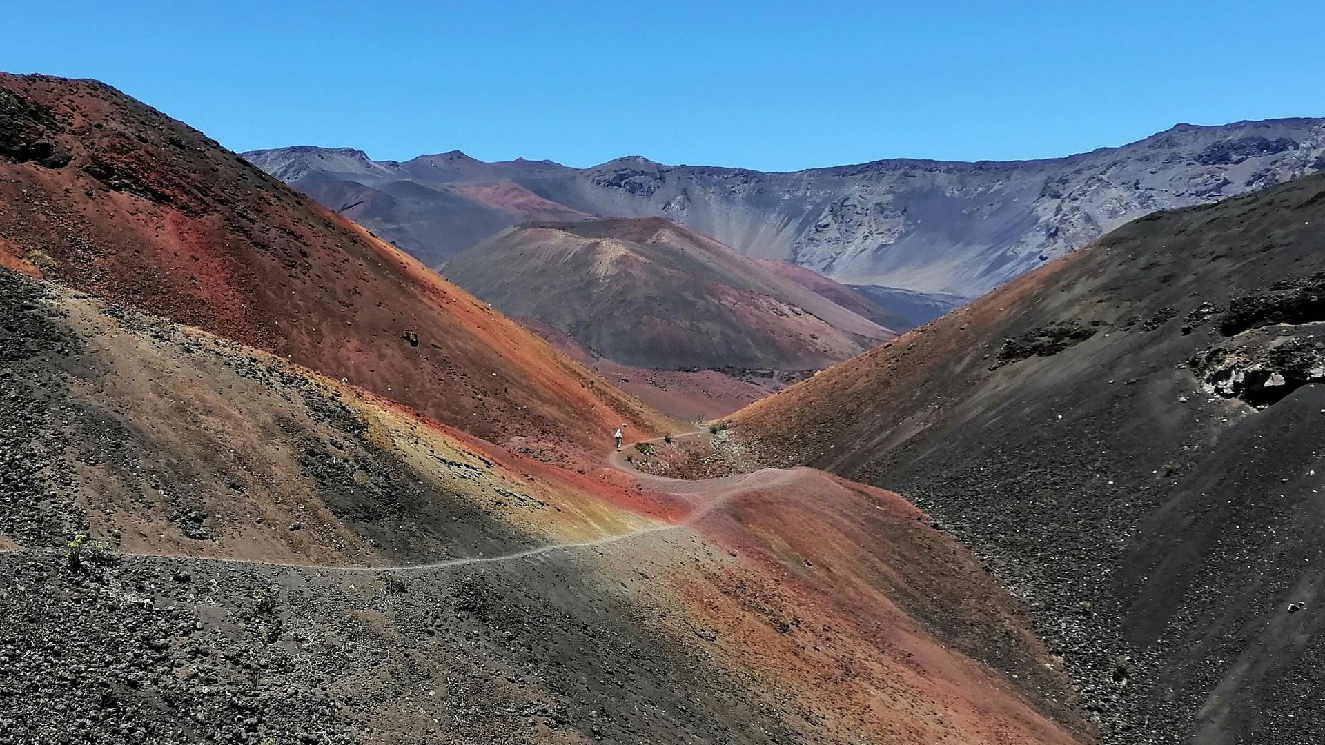 Colorful volcanic valley with a winding dirt path and distant mountains under clear sky.