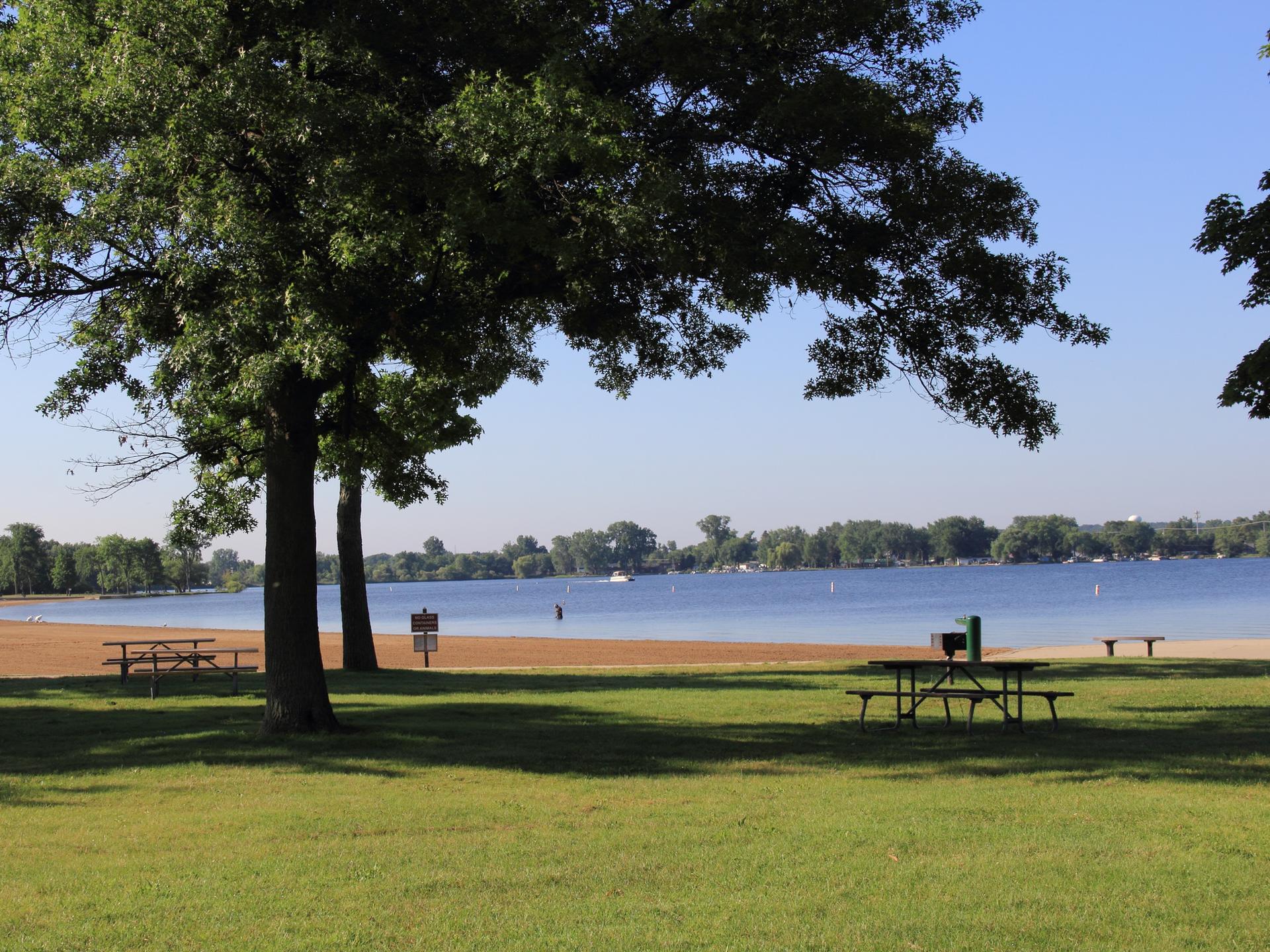 Sunny park with large trees, picnic tables, sandy beach, and a calm lake.