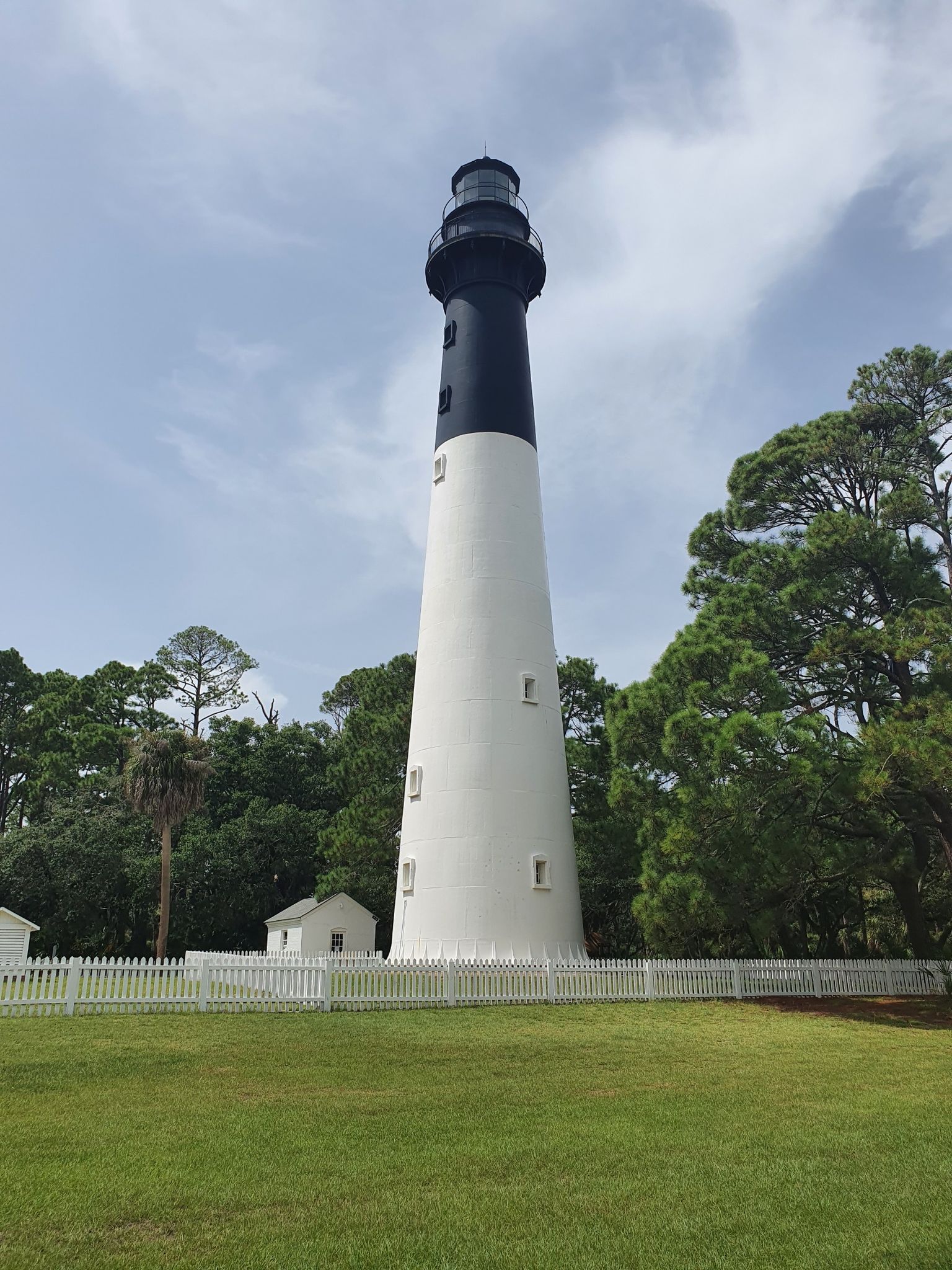White and black lighthouse on a sunny day, surrounded by green trees. - Hunting Island Lighthouse Beach Trail