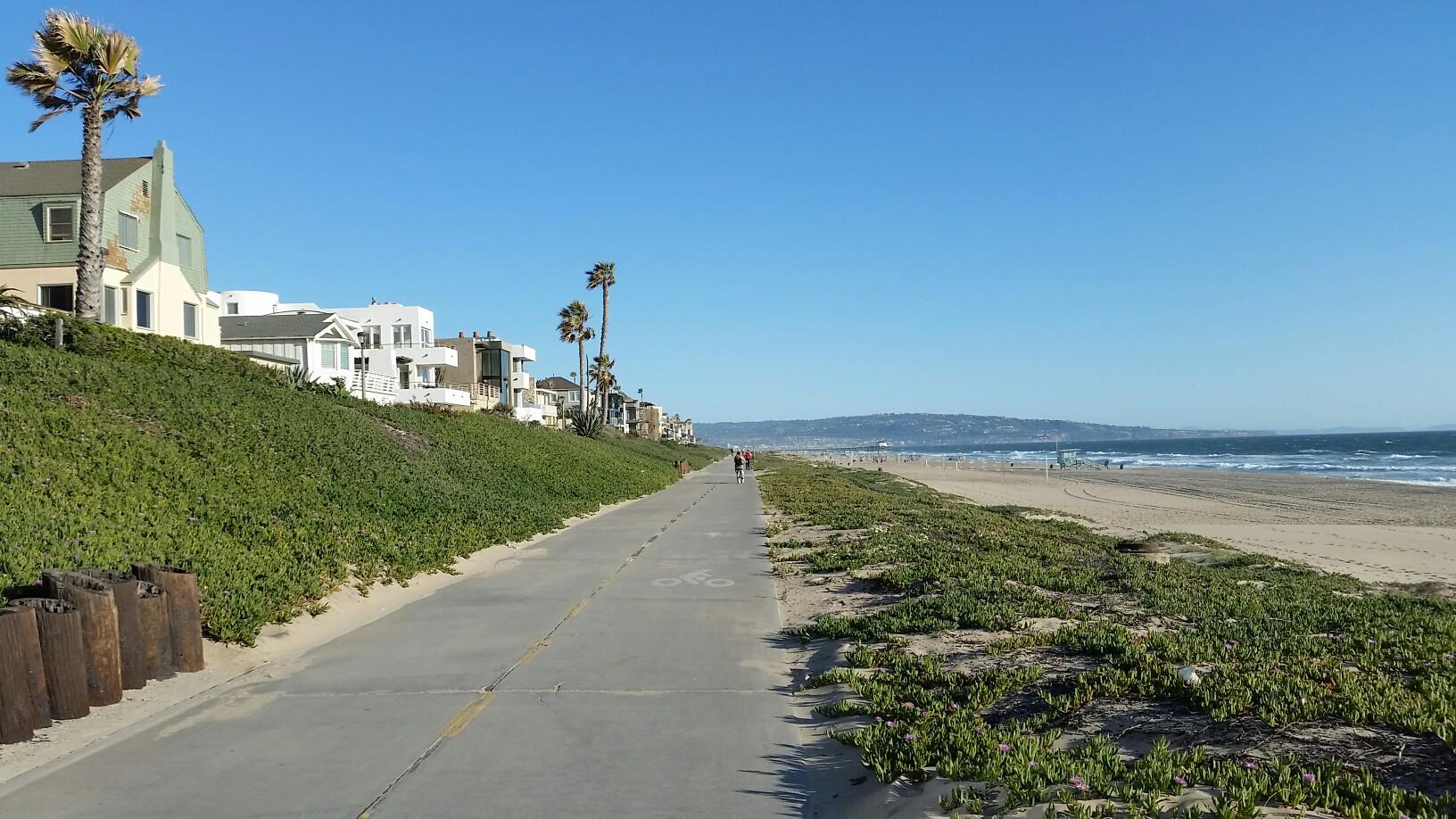 Sunny beach path with houses, green dunes, and distant ocean.