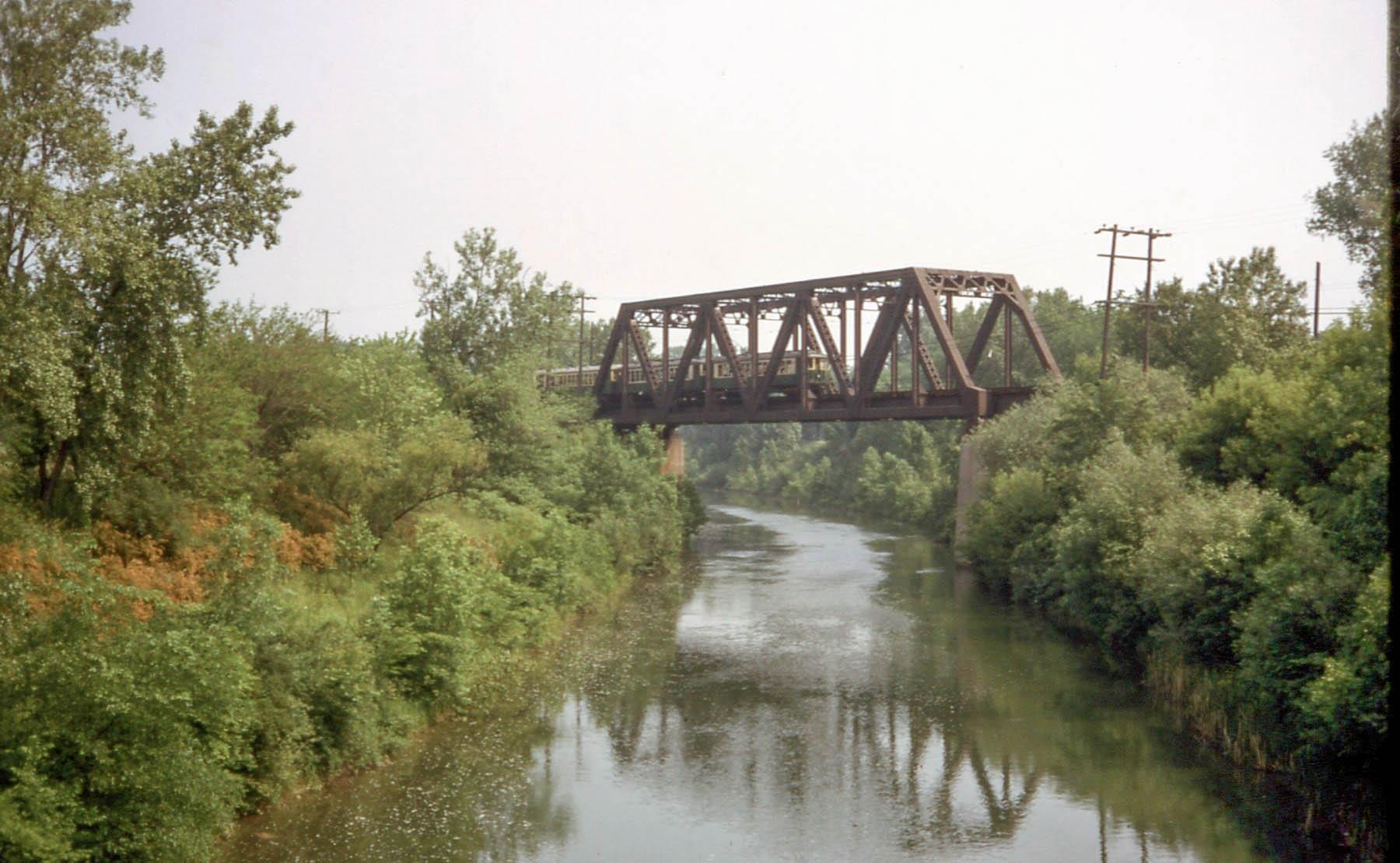 A metal truss bridge spans a narrow river surrounded by dense green trees.