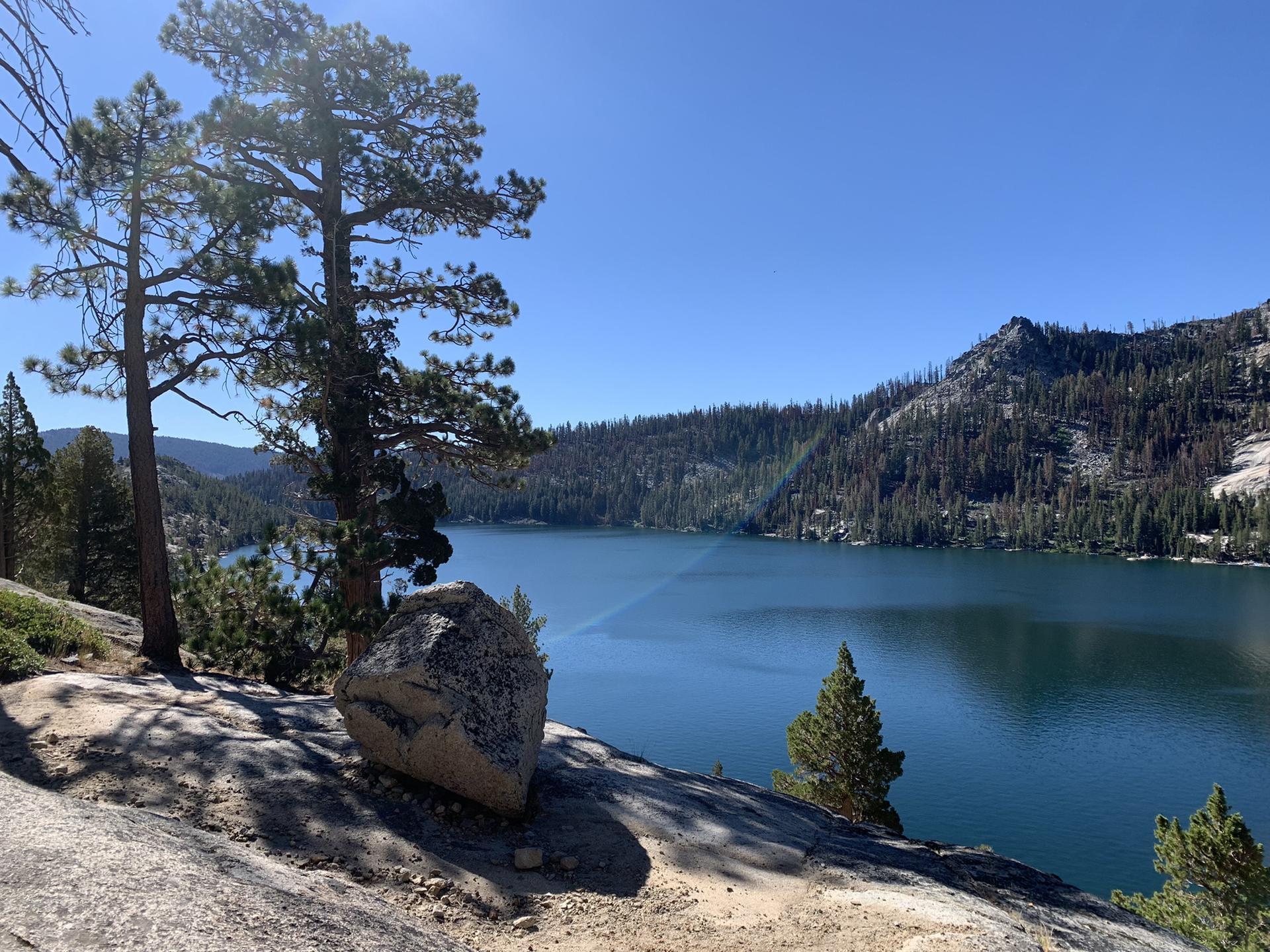 Sunny view of a clear blue lake surrounded by forested mountains and rocks.