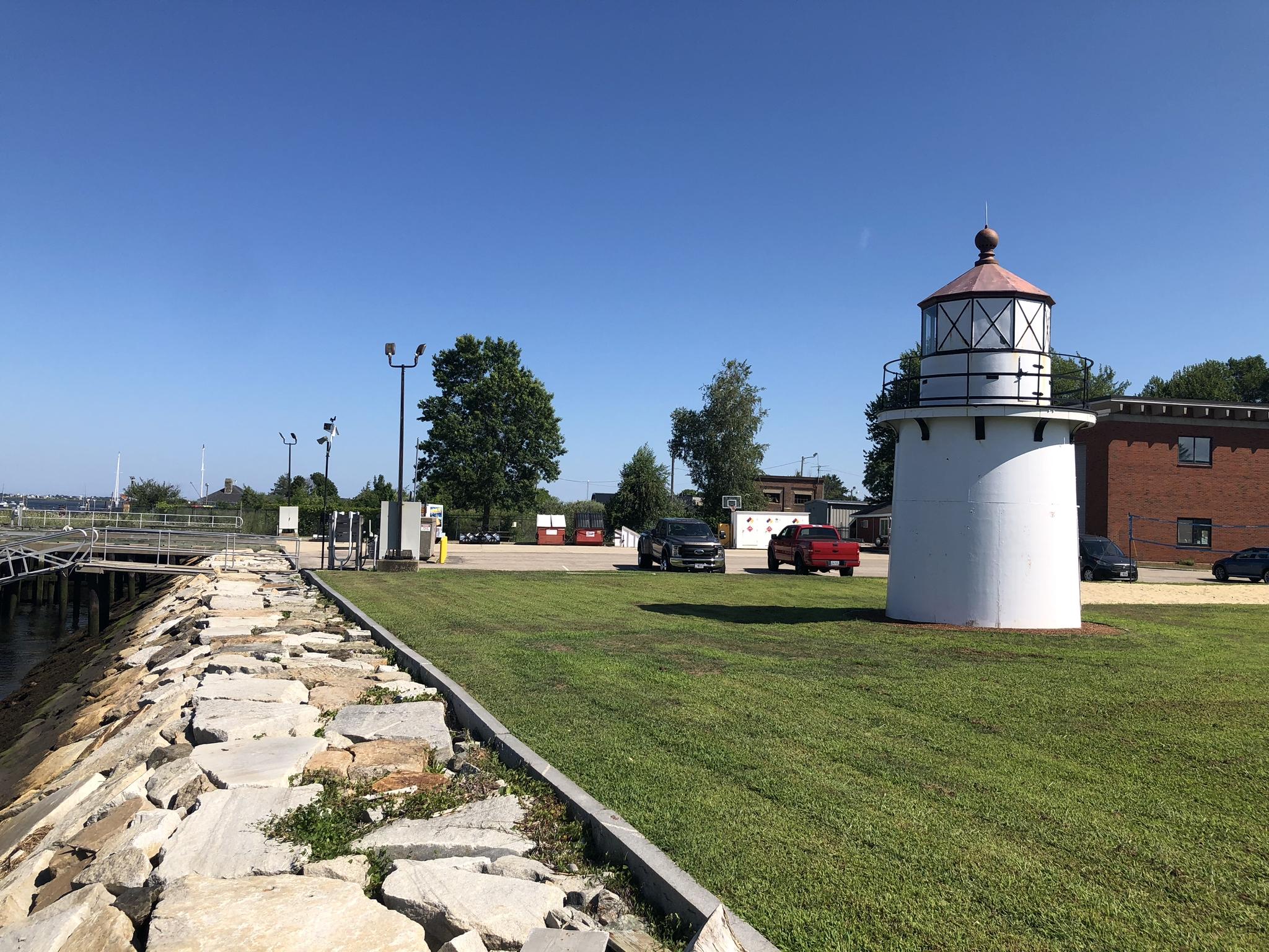 White lighthouse next to a grassy area with cars under a clear blue sky.