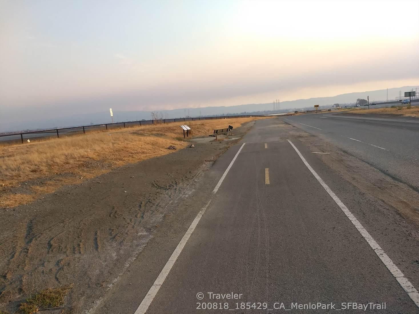 Paved path runs alongside a road through dry, hazy landscape.