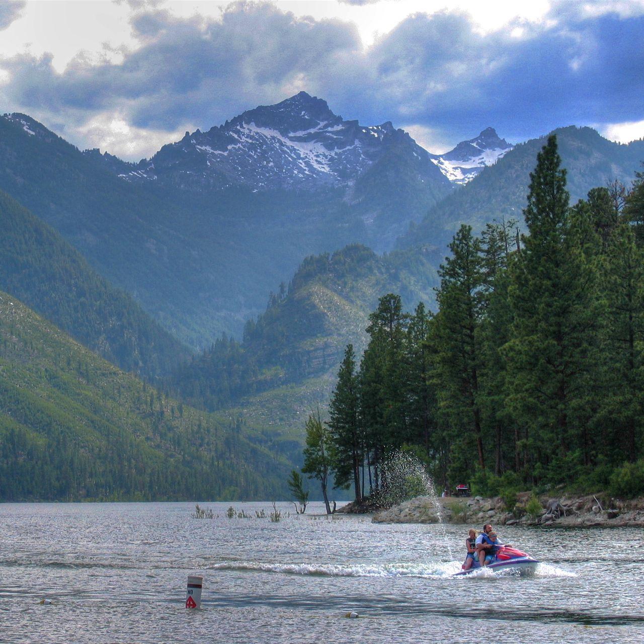 People jet skiing on a lake, surrounded by forested mountains with snowy peaks.