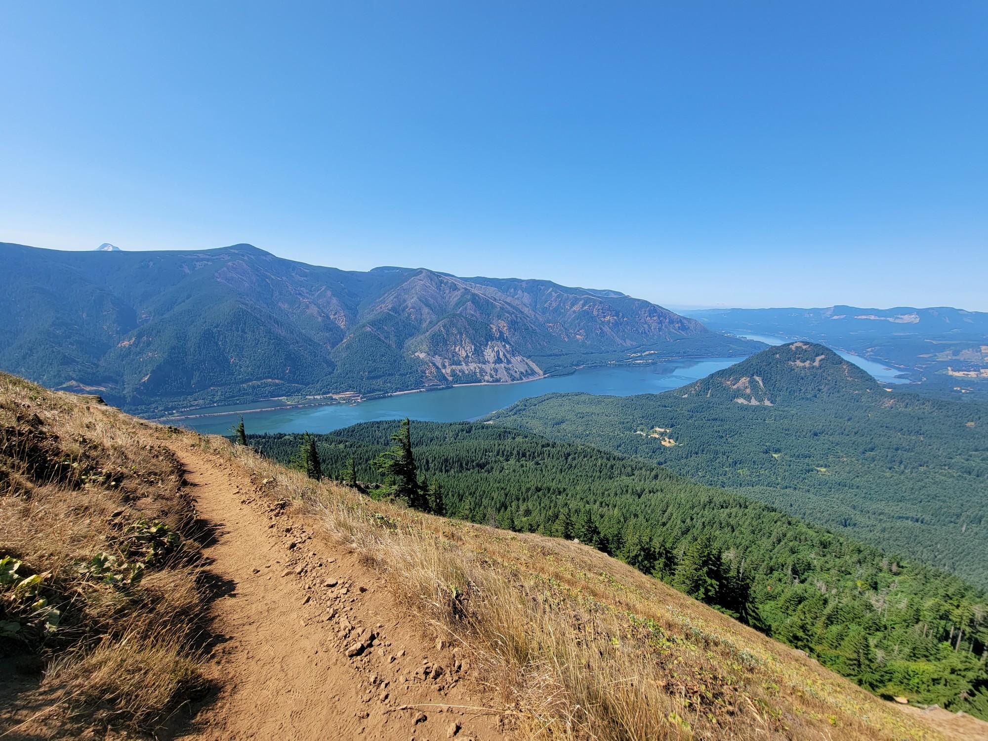 Dirt trail on a sunny hillside overlooking a wide river and distant mountains.