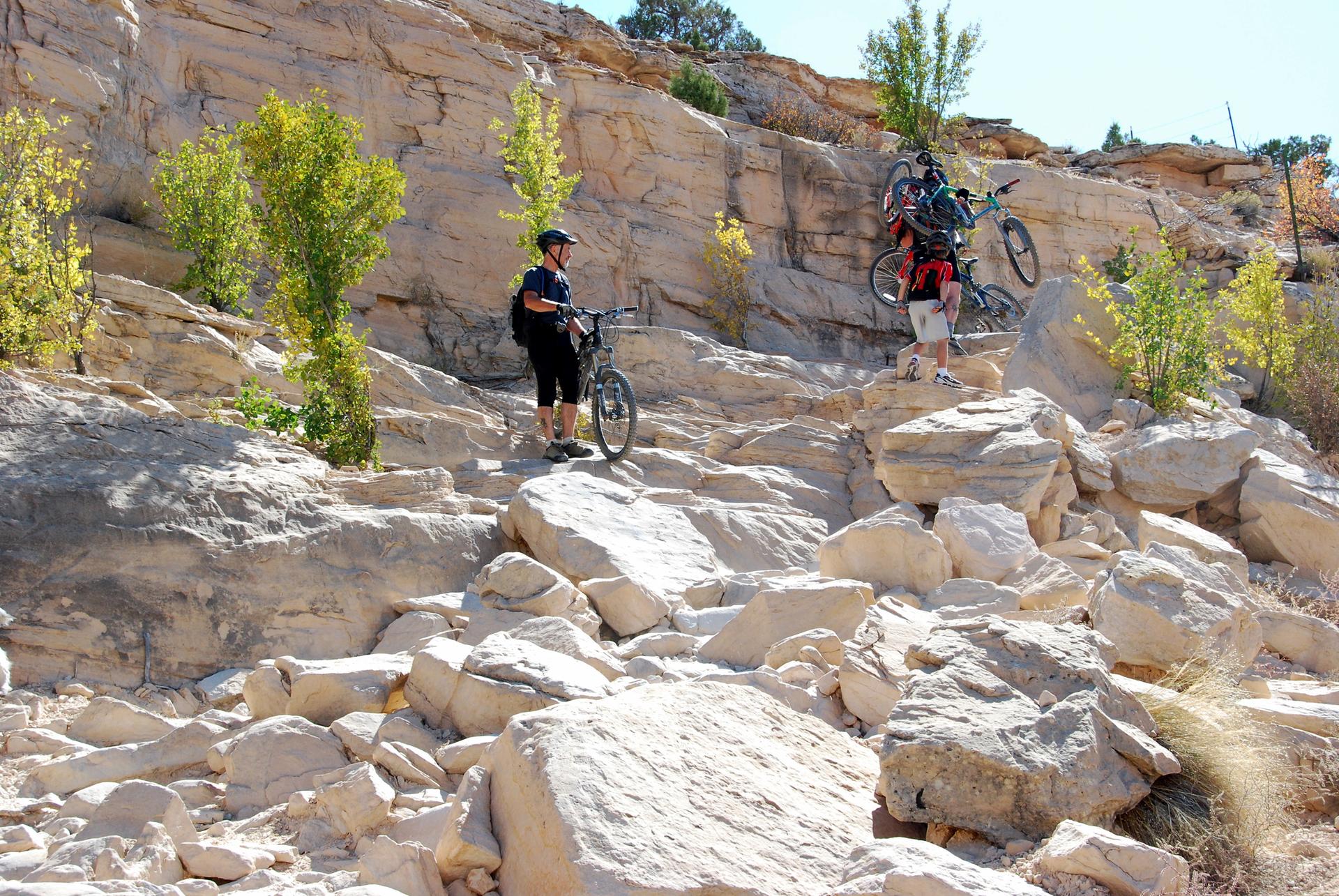 Two mountain bikers navigate a very rocky, steep path, one carrying their bike.