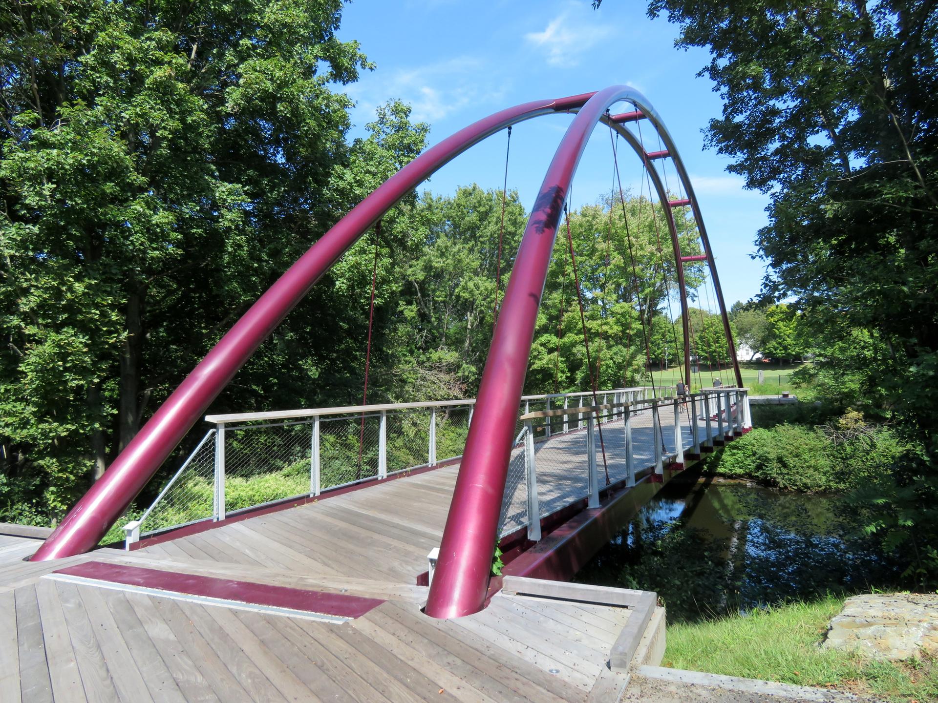 A red arched pedestrian bridge spans over a small stream on a sunny day.