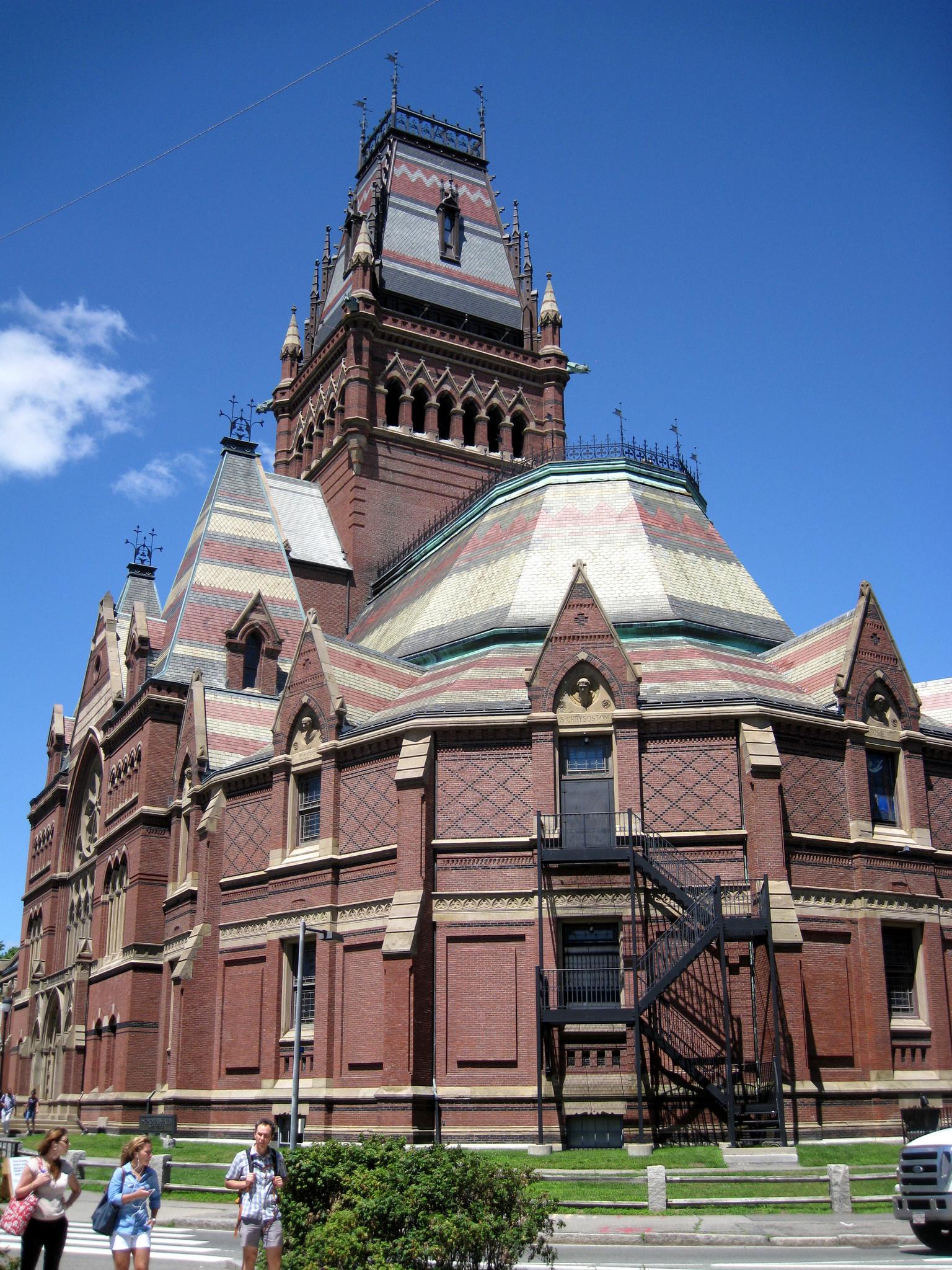 Ornate brick building with a tall tower under a bright blue sky. People walk nearby.