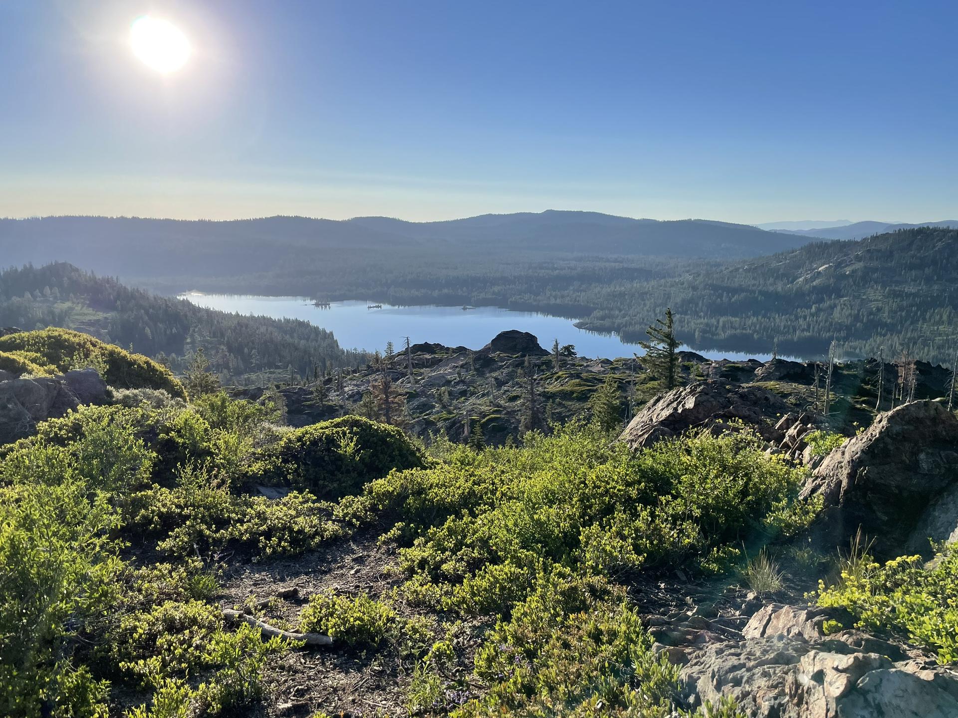 Bright sun over a lake surrounded by green hills and forests, clear sky.