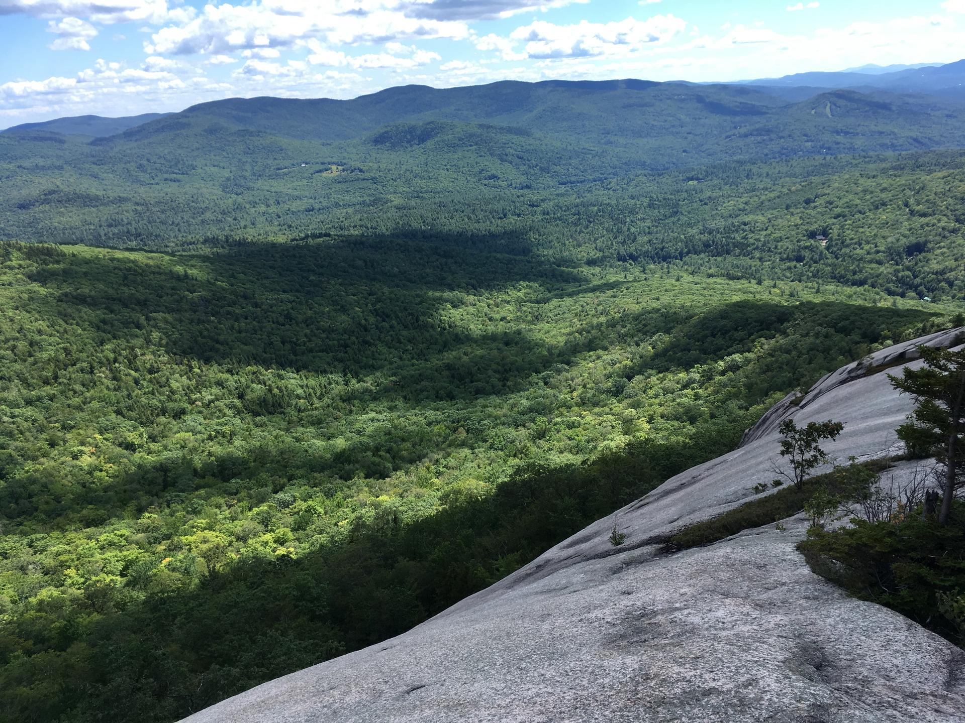 Steep rocky slope overlooking a vast green forest valley under a partly cloudy sky.