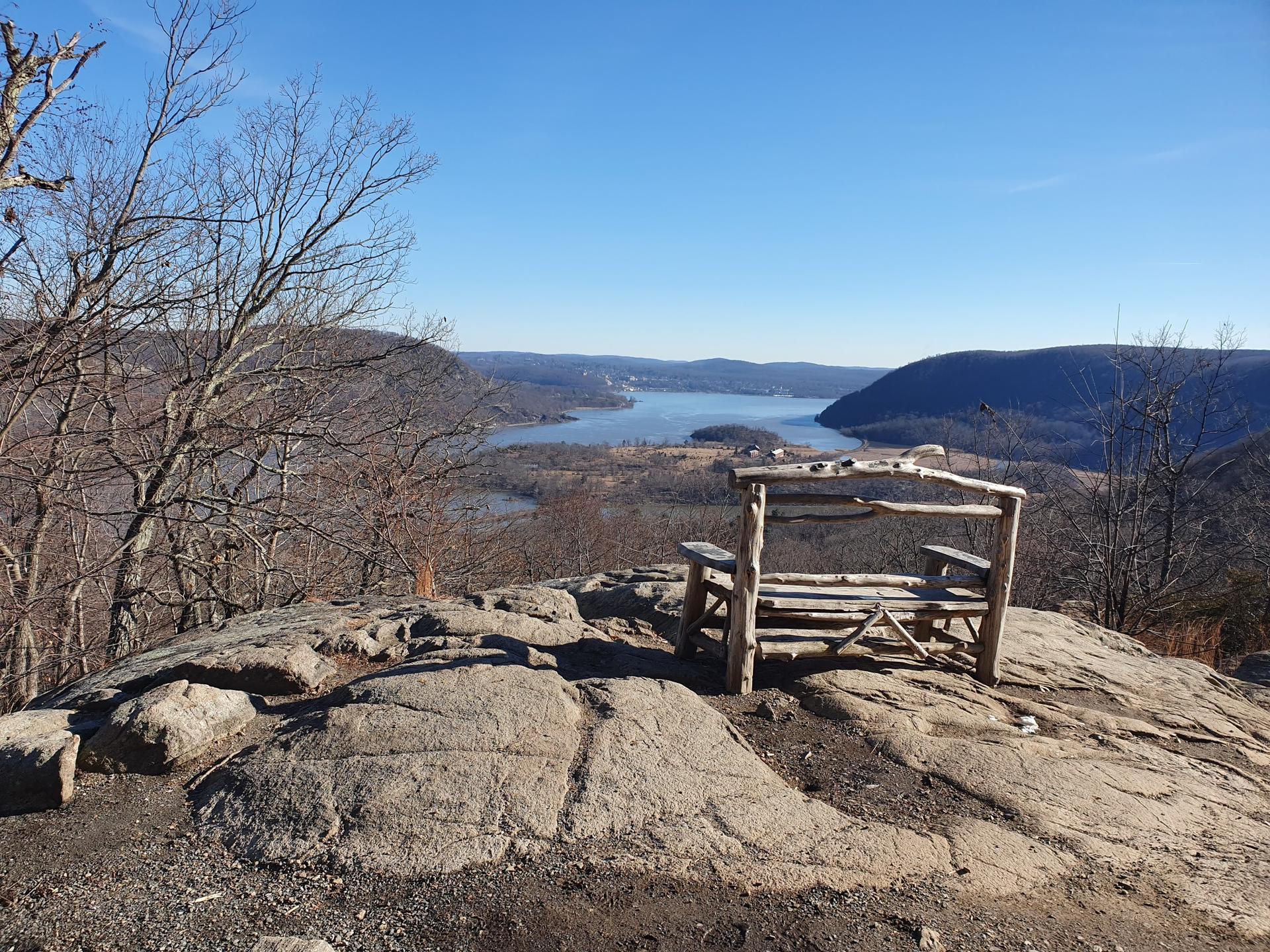 Wooden bench on rocky peak overlooking a wide river and distant hills.