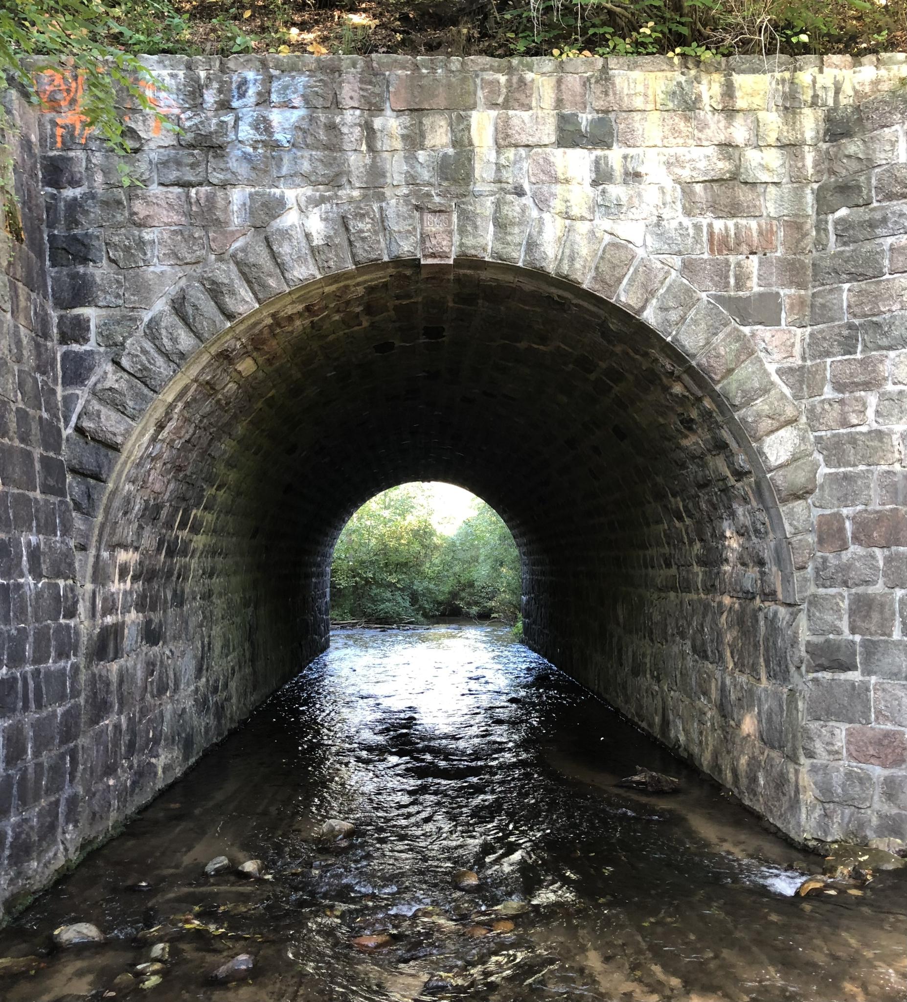 Stone archway tunnel with a stream flowing through, bright light at the end.