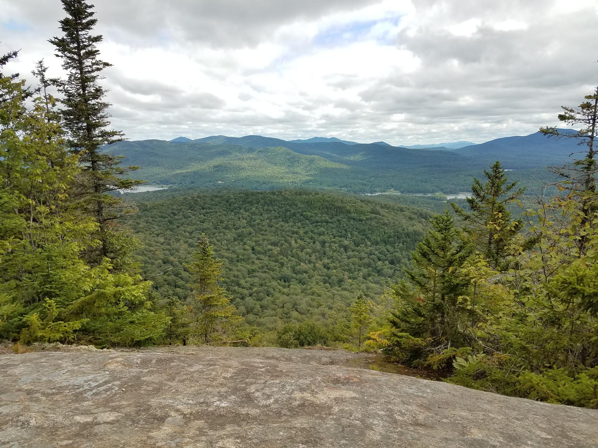 Overcast view from a rocky ledge overlooking a vast forest and distant mountains.