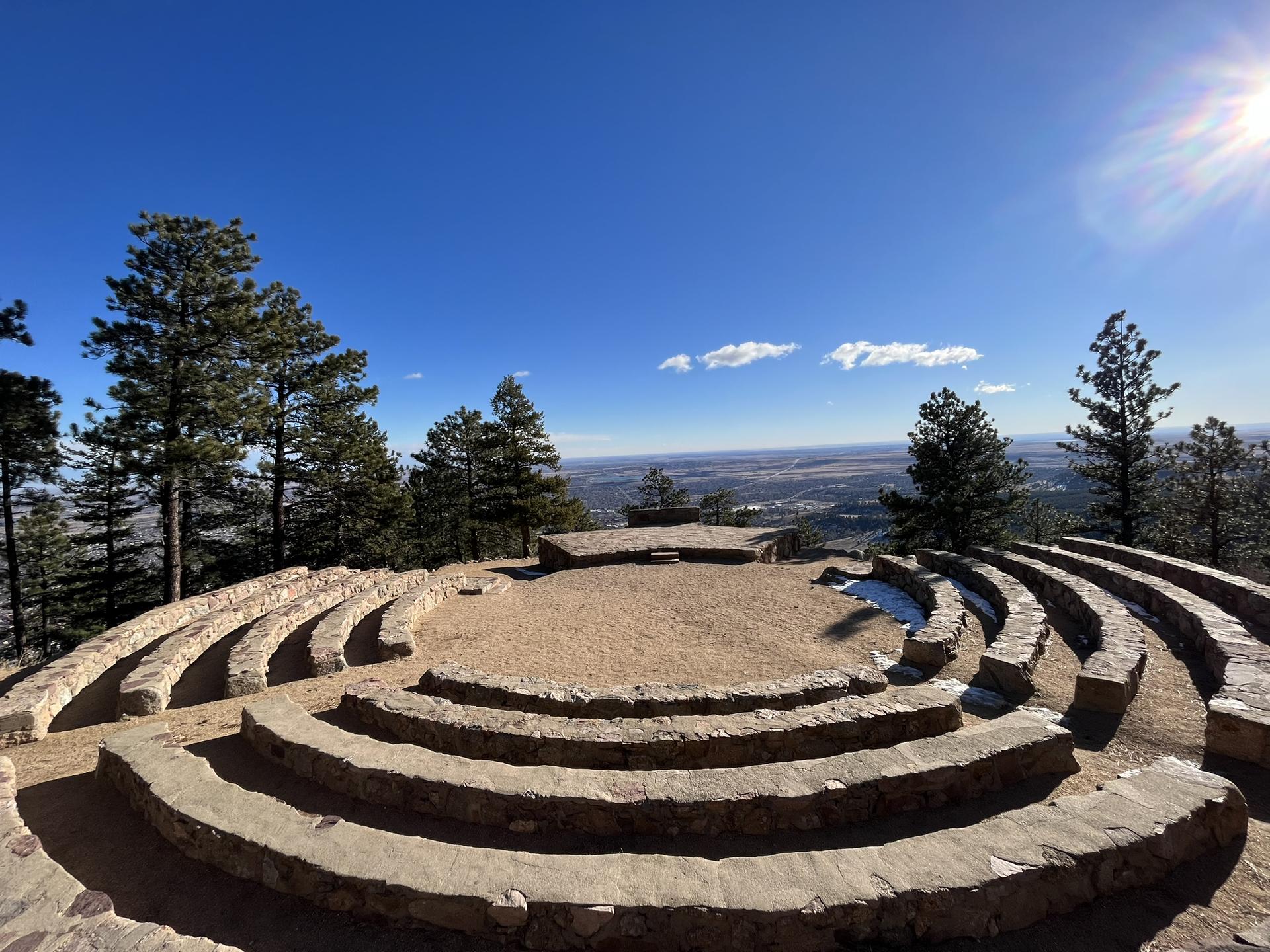 Stone amphitheater on a mountain top, surrounded by pine trees under a bright blue sky.