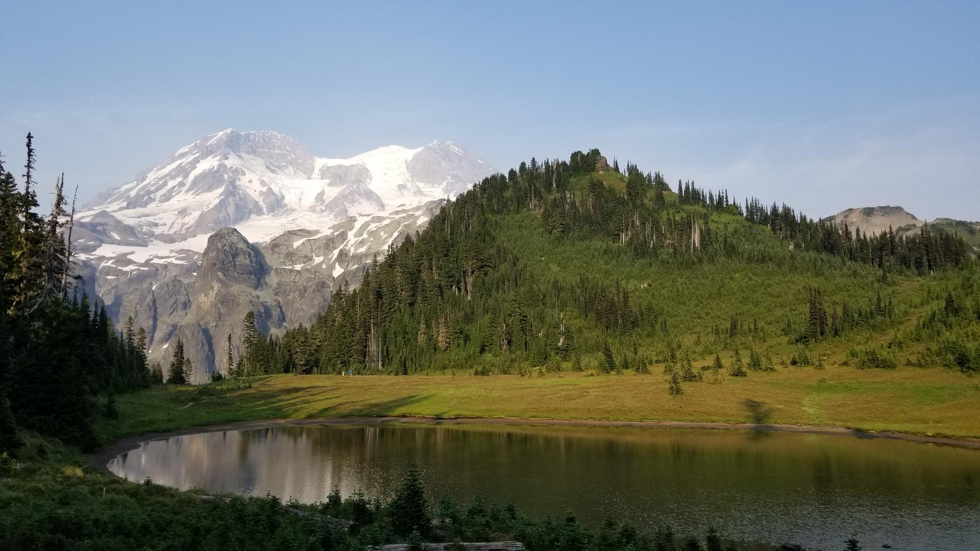 Snowy mountain peak, green forest, and calm lake reflecting the sky.