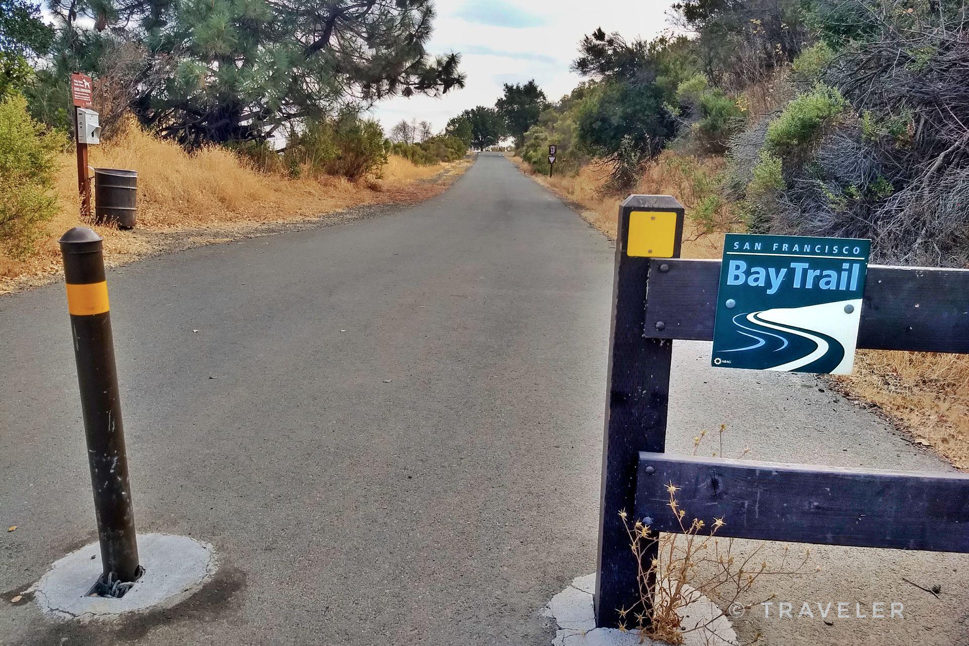 Sunny paved path entrance with a wooden barrier, bollard, and dry vegetation.