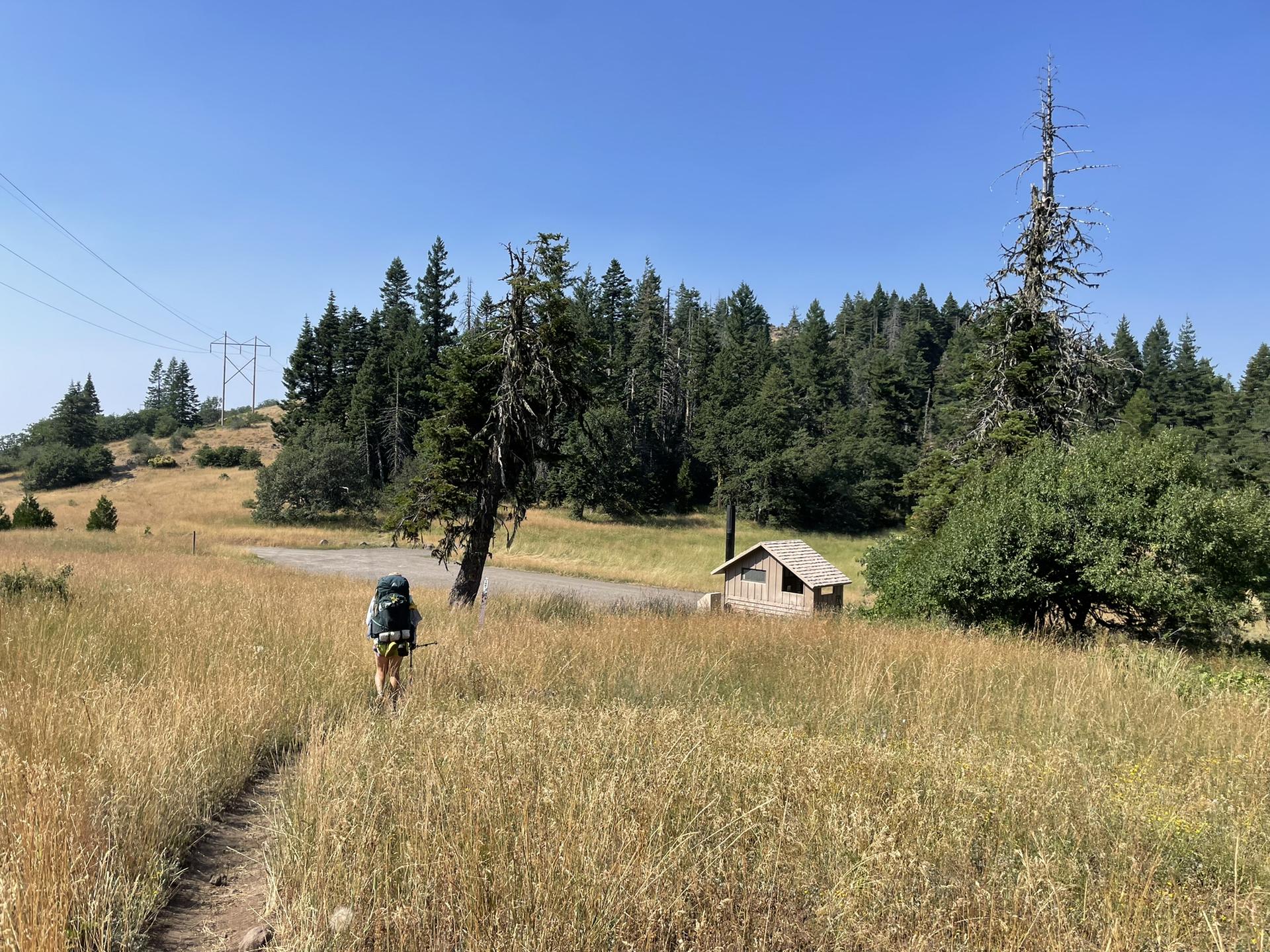 Hiker walks on a dirt path through a dry grassy field under a clear sky.