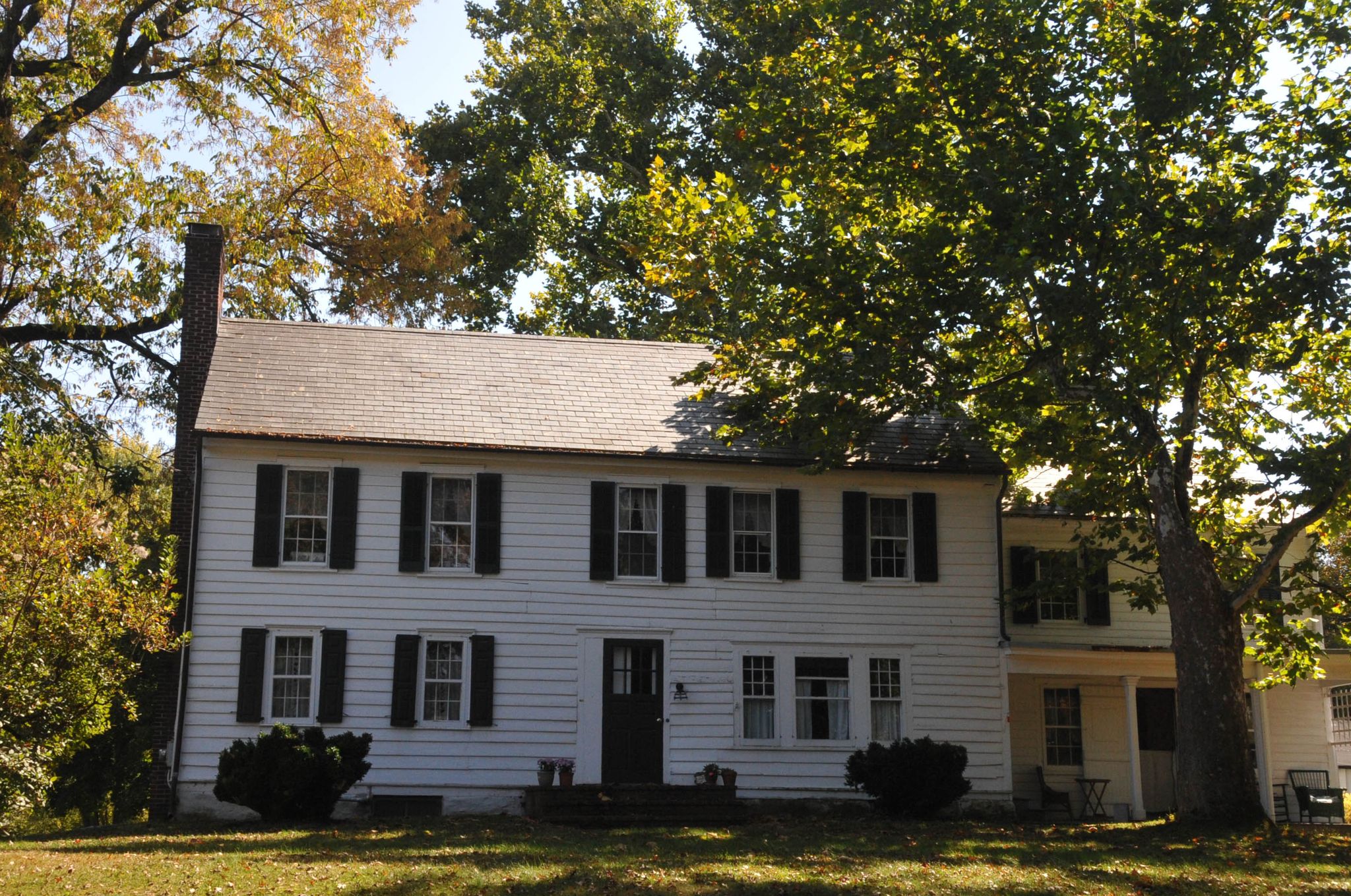 White two-story house with black shutters surrounded by trees on a sunny day. - Hopewell Loop