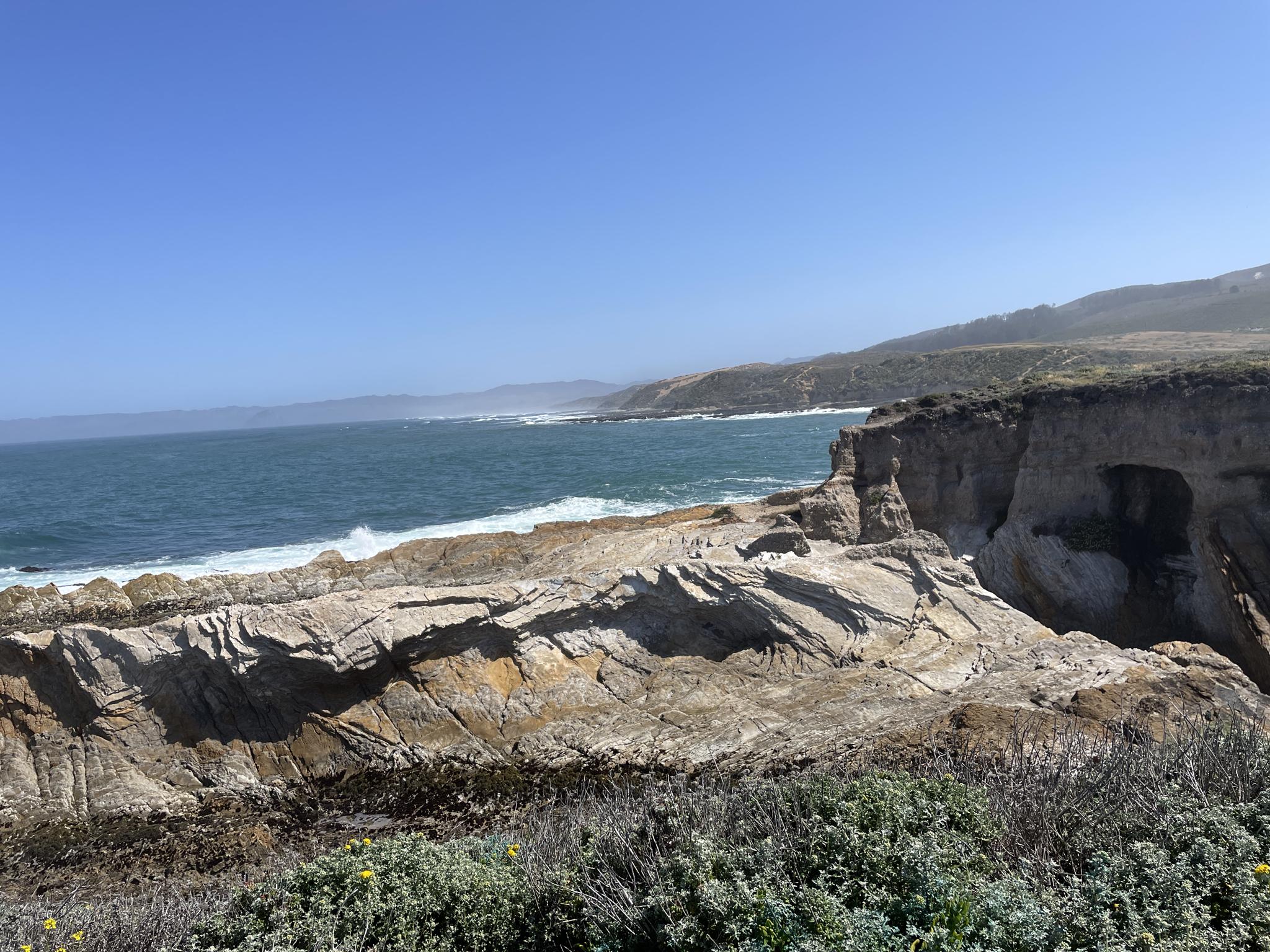 Sunny coastal cliffs and ocean with waves, green and brown vegetation.