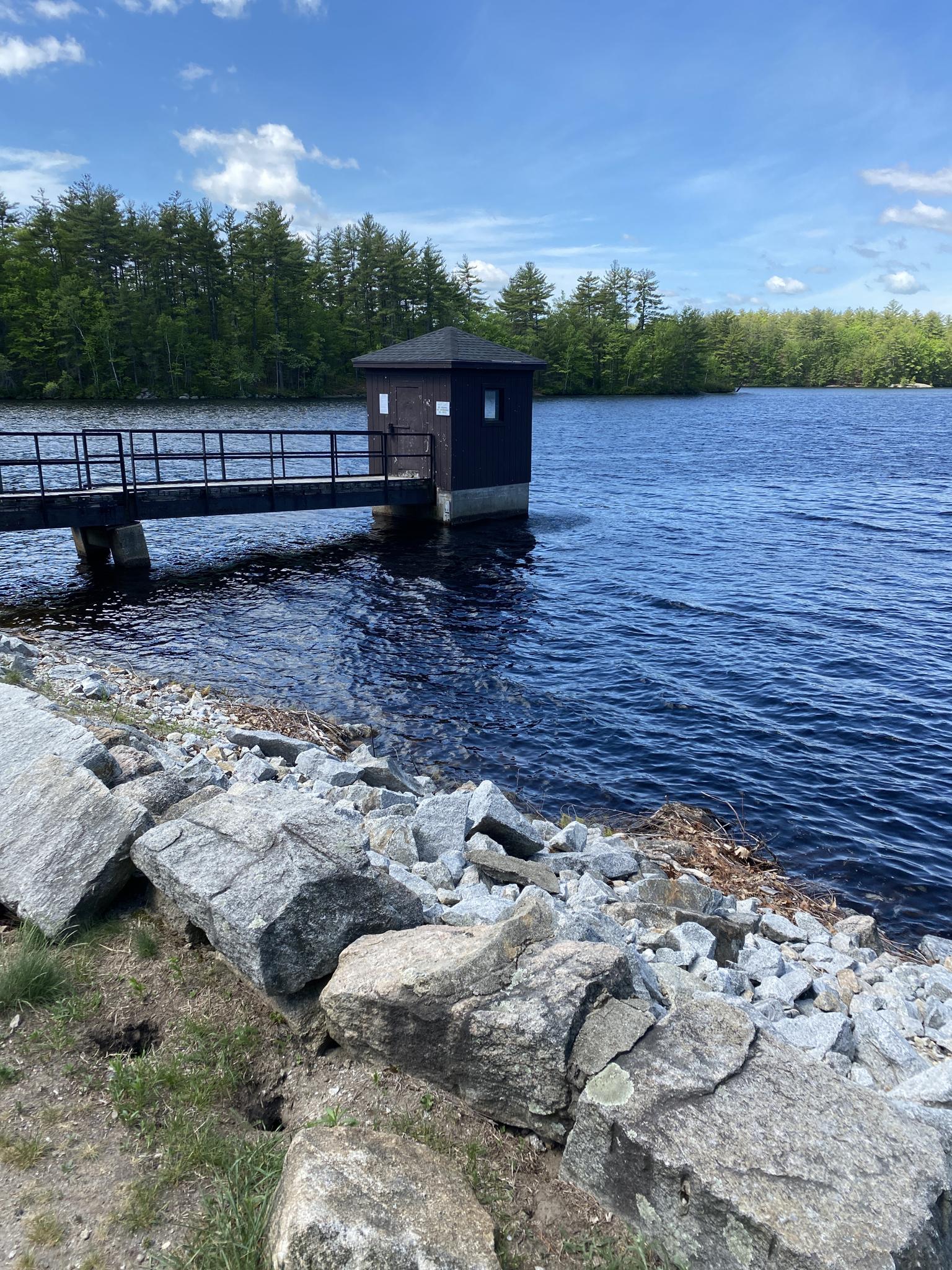 A small dark building on a lake with a bridge under a blue sky.