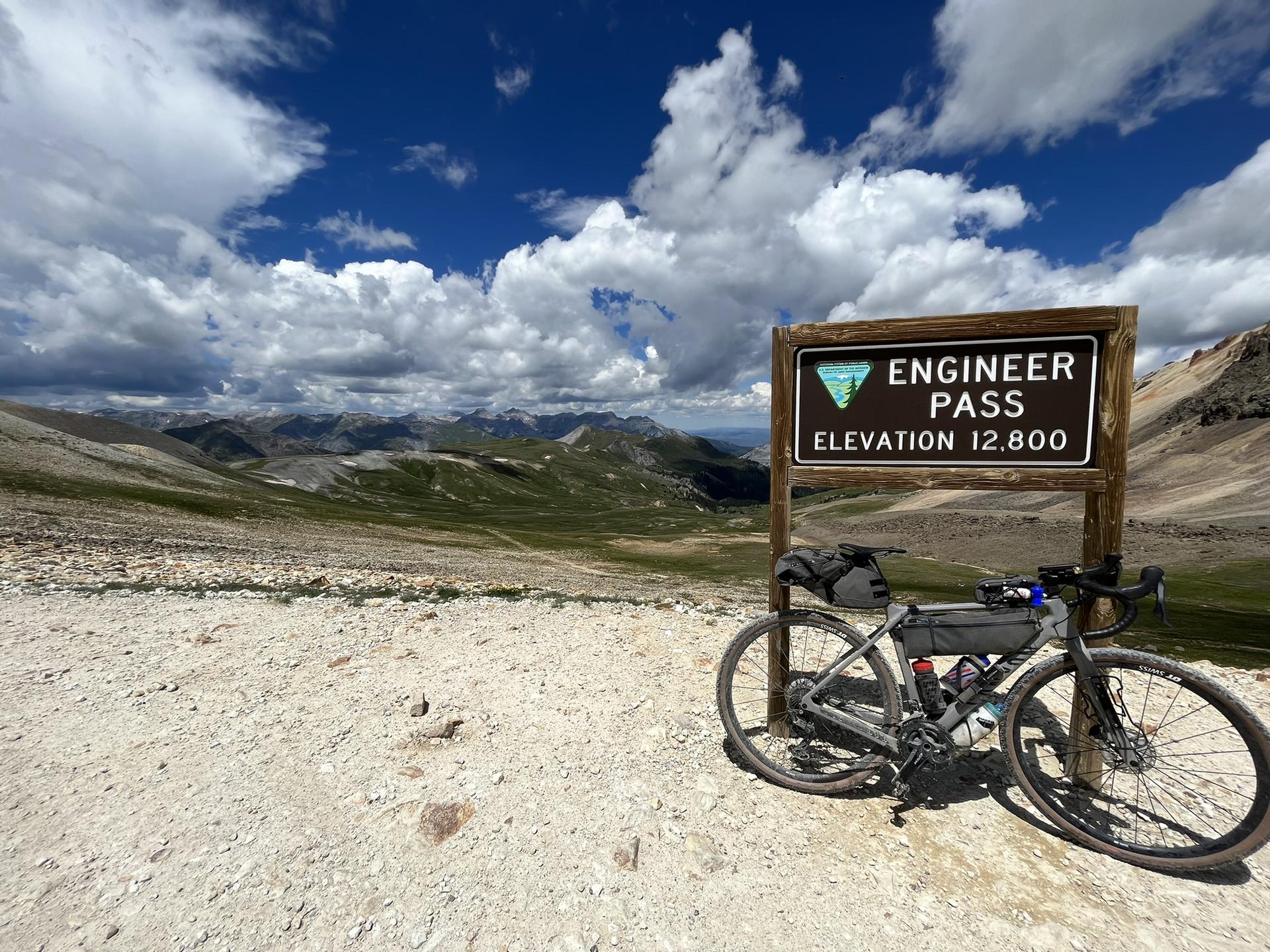 Bicycle parked by a pass sign, overlooking vast mountains and cloudy sky.