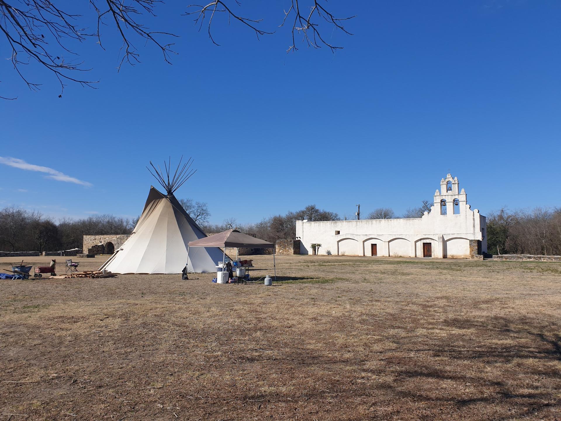 Large teepee and white building in a grassy field under clear blue sky.