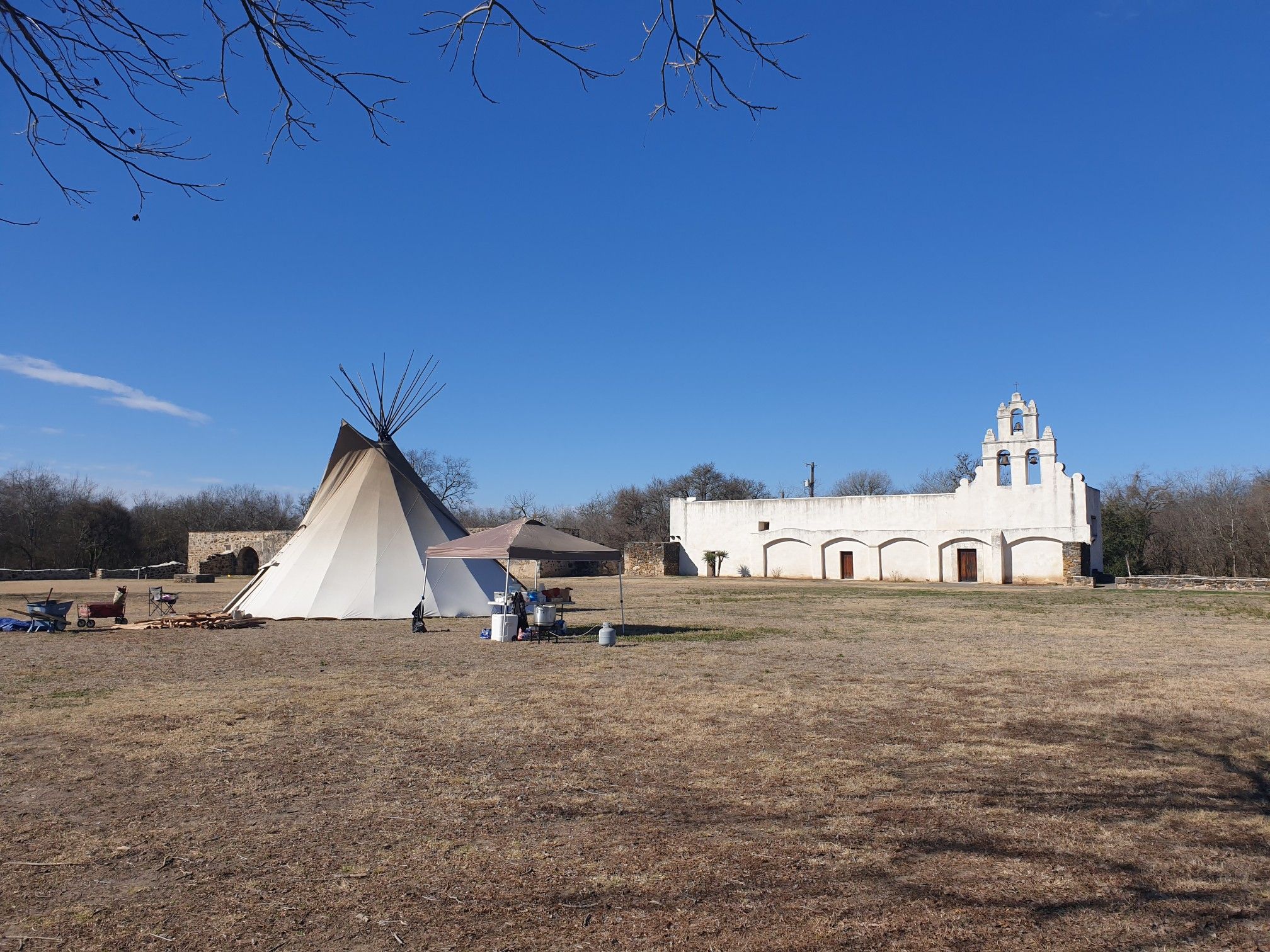 Large teepee and white building in a grassy field under clear blue sky. - River Walk Mission Reach
