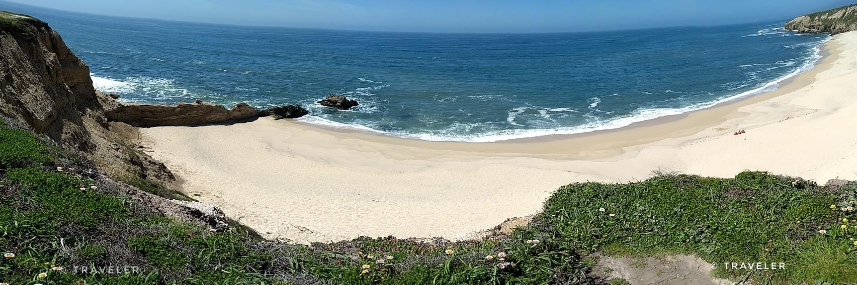Panoramic view of a secluded sandy beach with blue ocean and green cliffs.