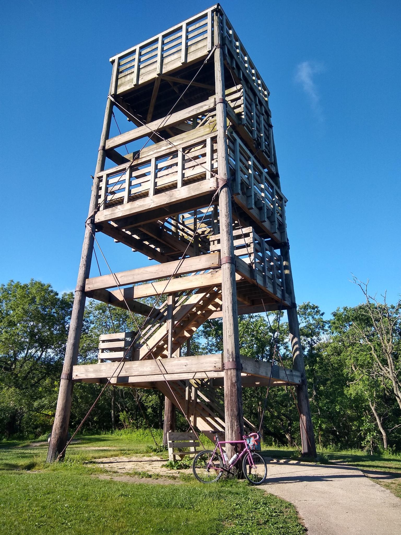 A tall wooden observation tower stands under a clear blue sky, with a bicycle.