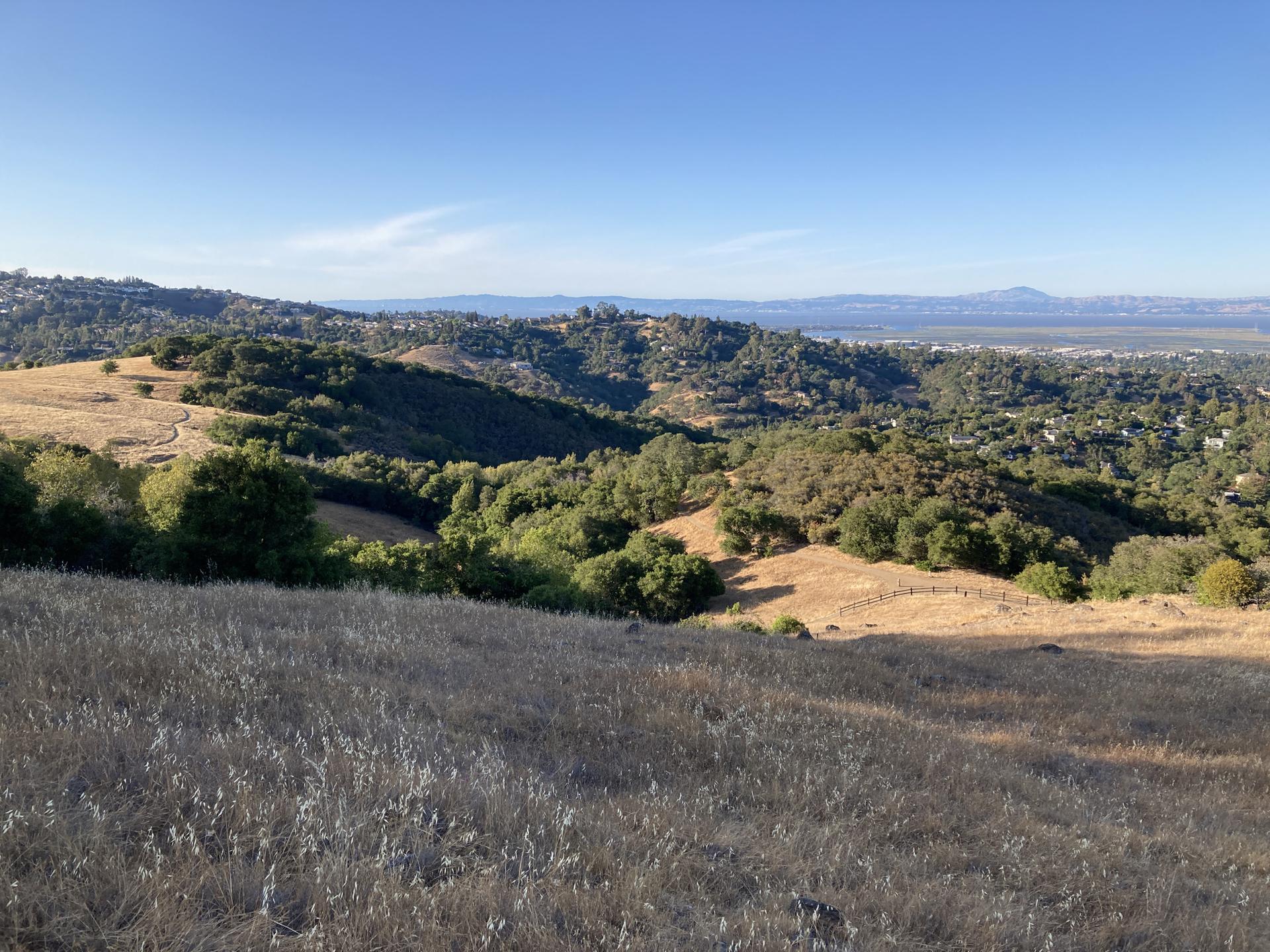 Golden grassy hills overlooking a green valley and distant water under clear sky.