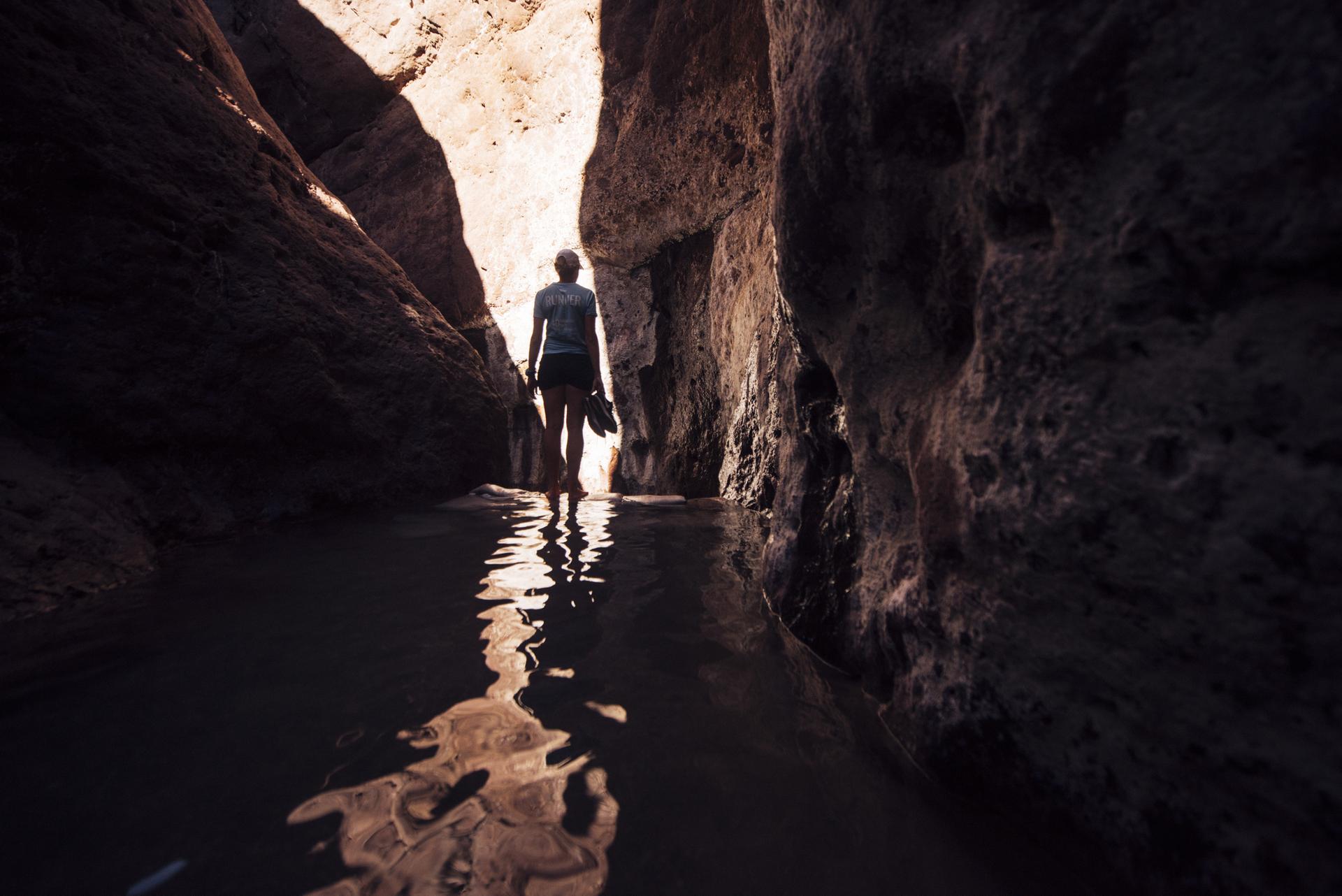 Person wading through water in a dark, narrow slot canyon, light ahead.