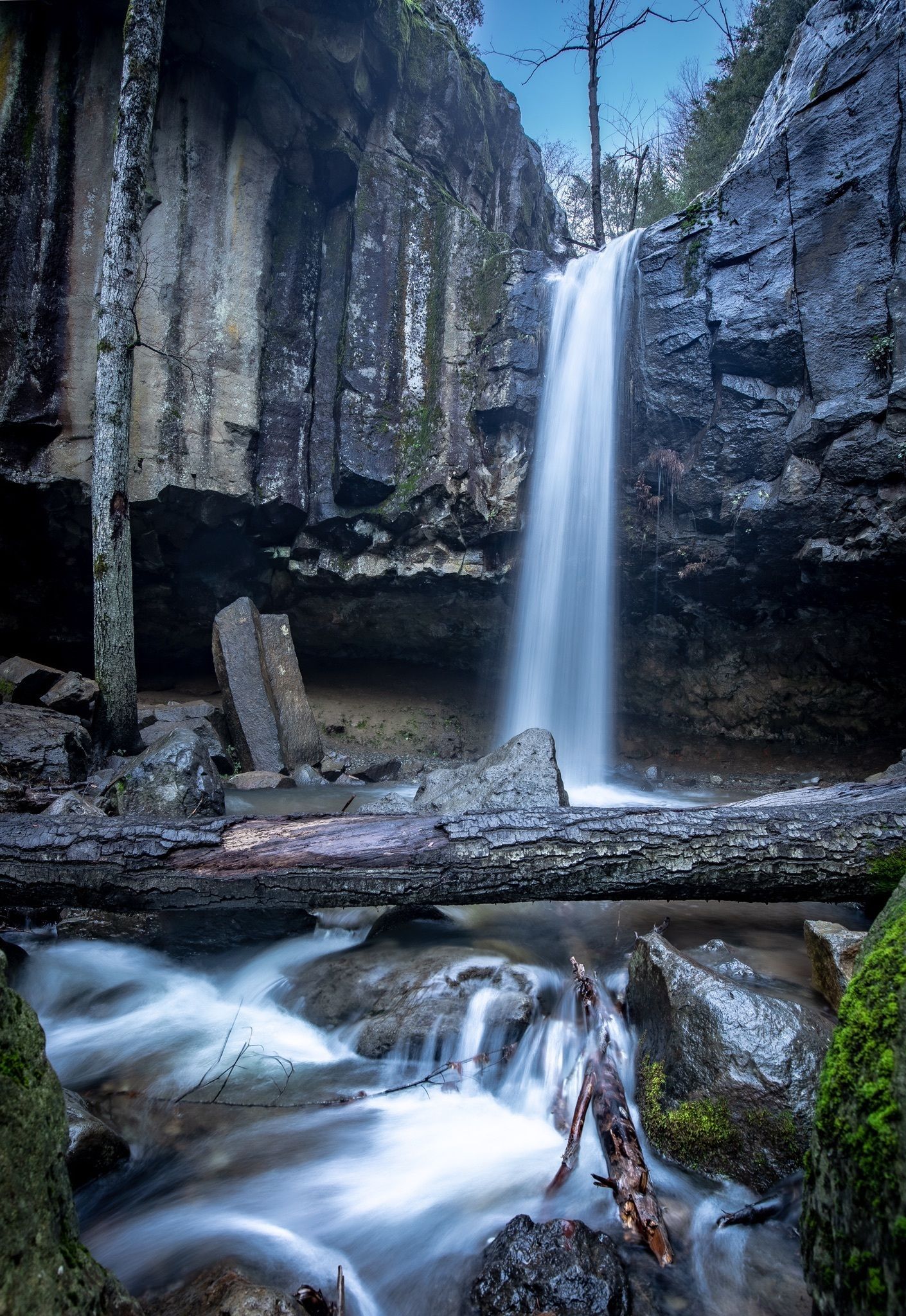 Soft light illuminates a waterfall, rocky cliffs, and a stream with a log. - Hedge Creek Falls Trail