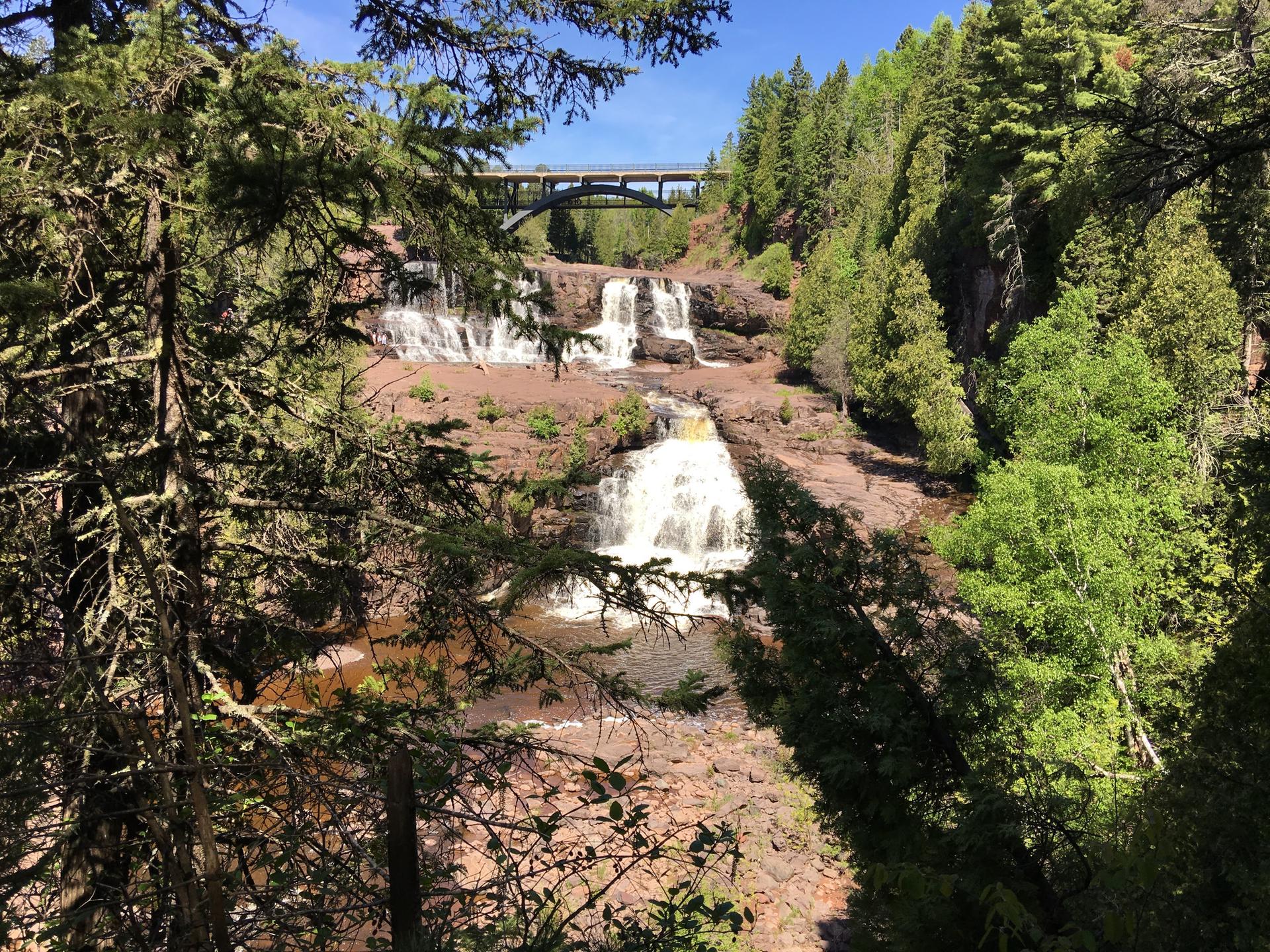 Sunny view of a multi-tiered waterfall flowing over red rocks, surrounded by green trees.