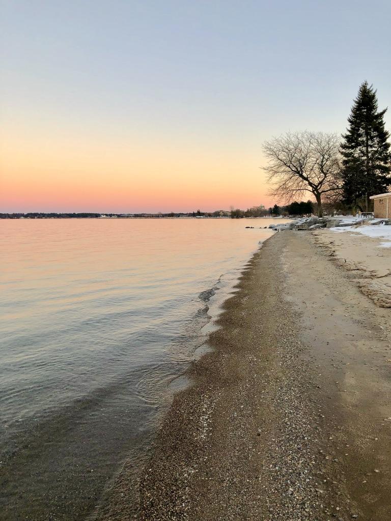 Serene shoreline at sunset with calm water, sand, and distant trees.