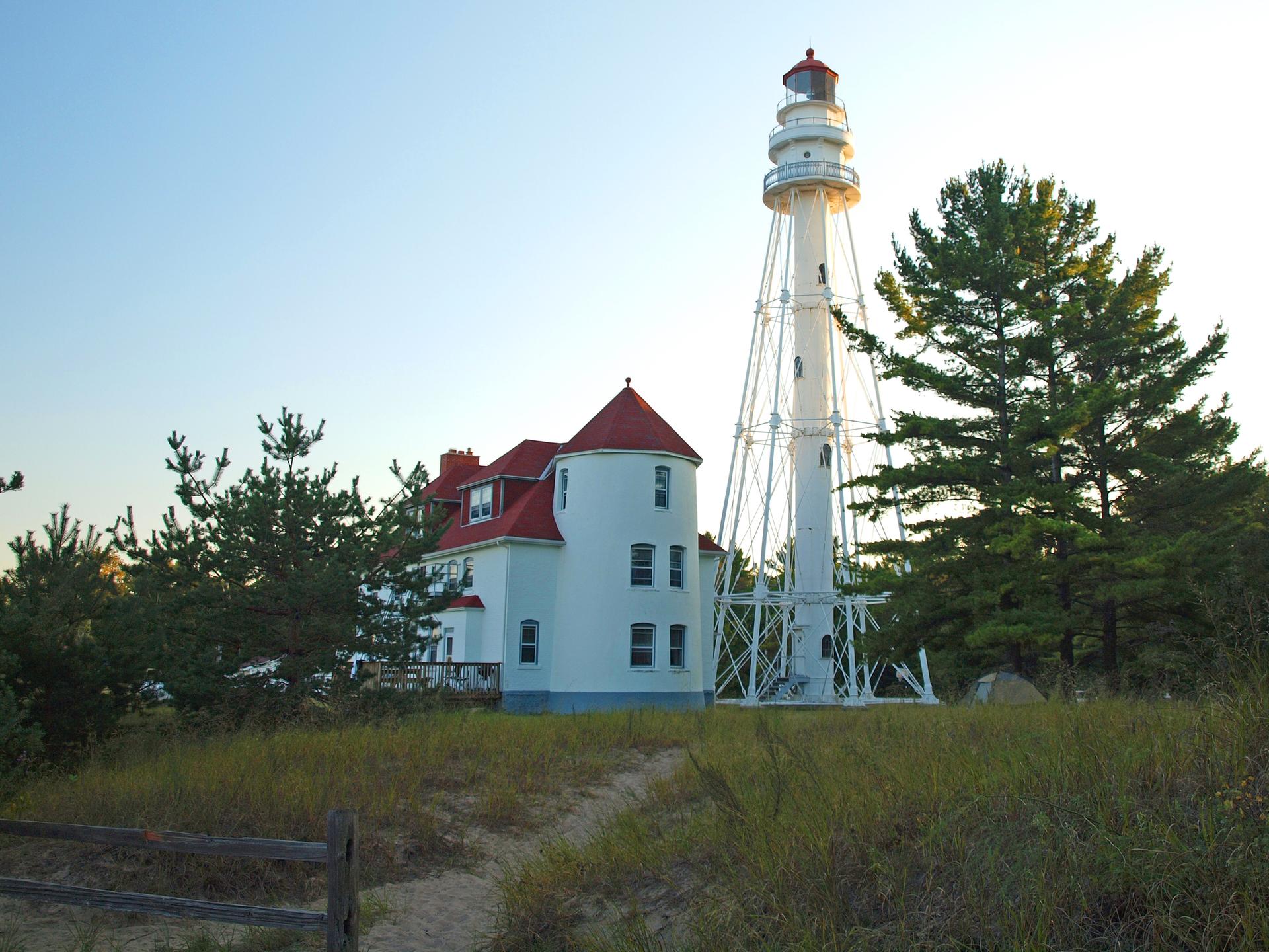 A white lighthouse and building with a red roof stand by trees at sunset.