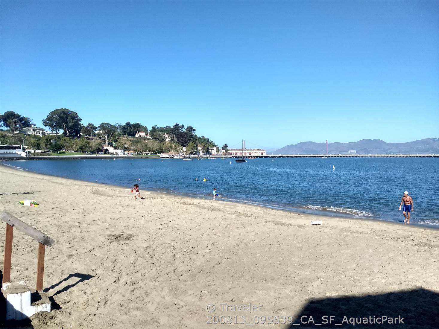 Sandy beach with calm water, distant bridge, children playing, bright sun.