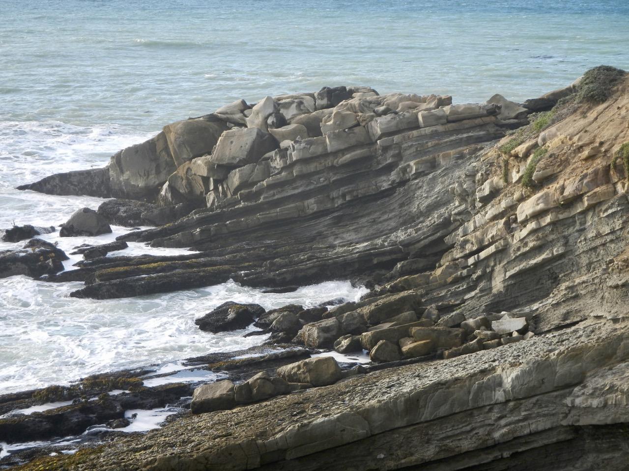 Layered rock formations on a rugged coastline with waves crashing.