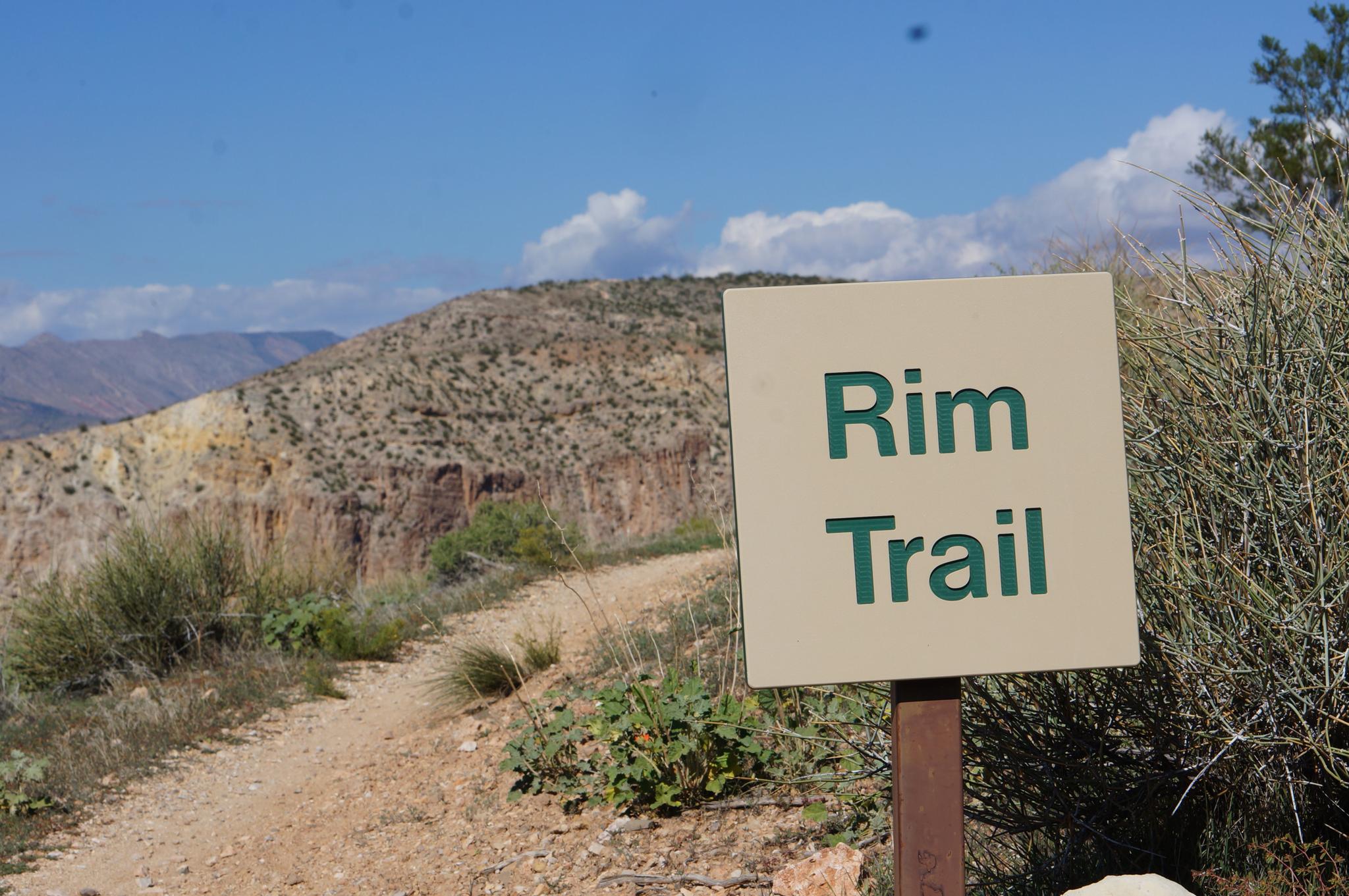 A 'Rim Trail' sign next to a dirt path in arid, sunny hills.
