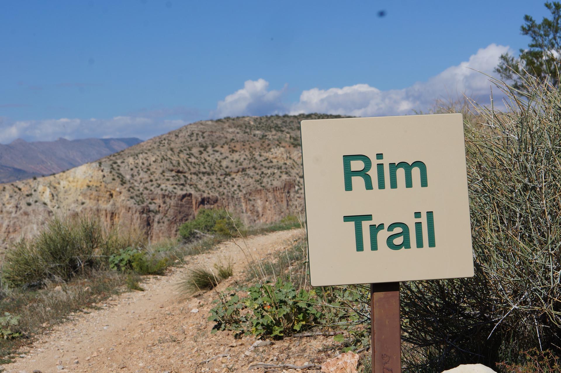 A 'Rim Trail' sign next to a dirt path in arid, sunny hills.