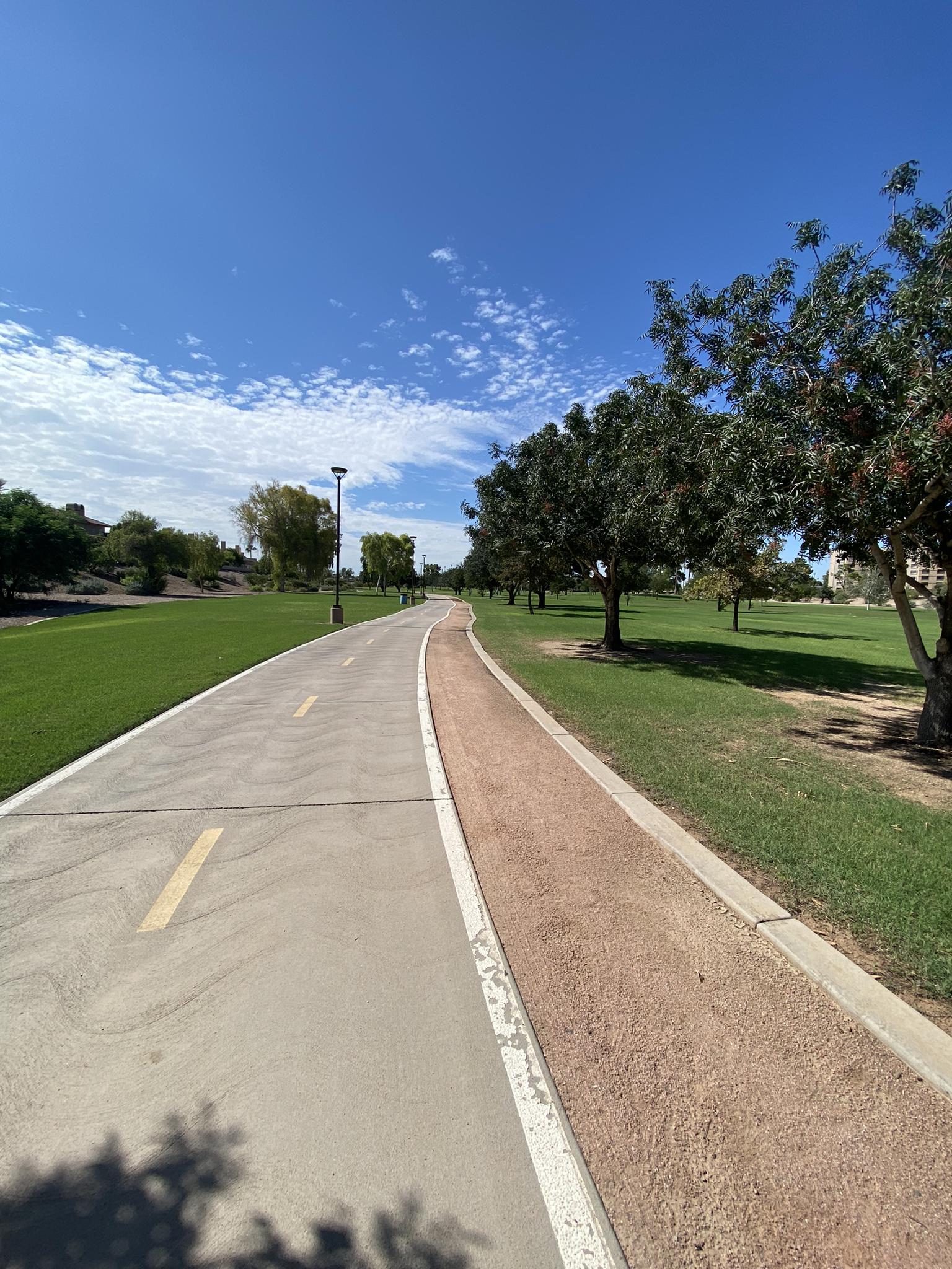 A paved and dirt path winding through green grass under a blue sky.