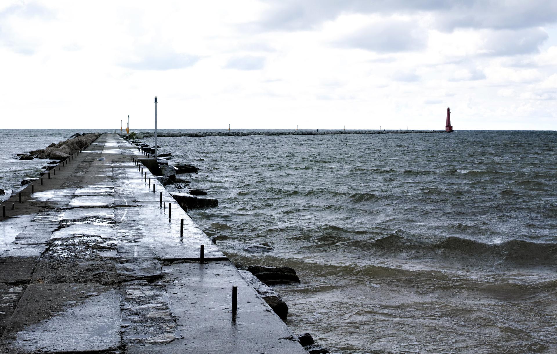 Wet concrete pier extends into choppy grey water with a distant red lighthouse.