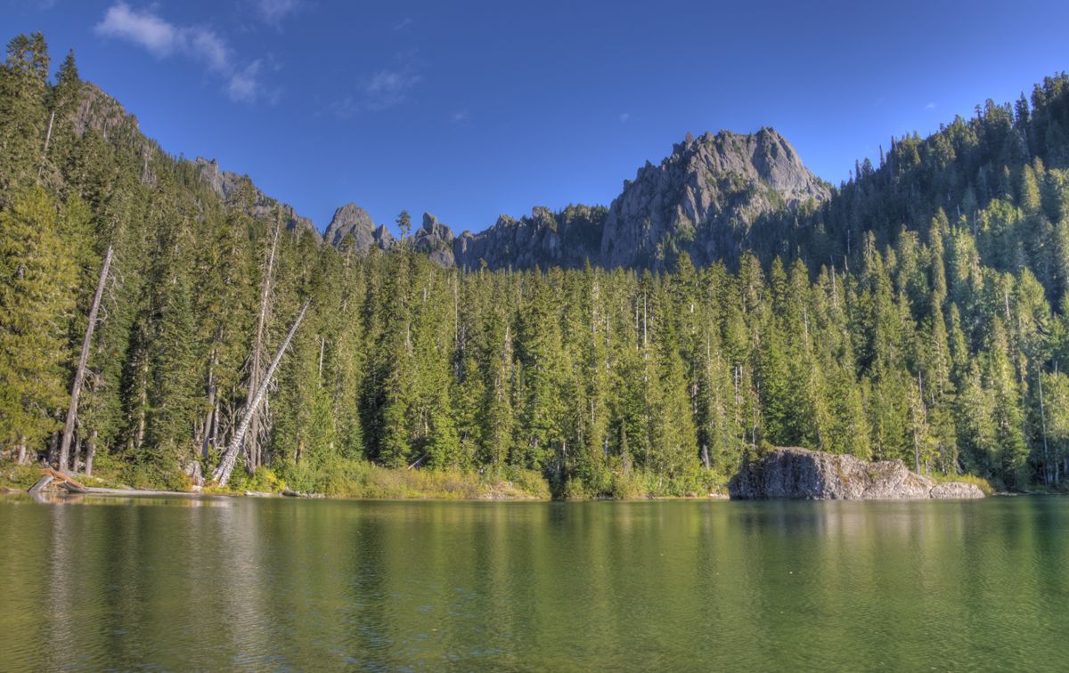 Calm lake reflecting dense forest and rugged mountains under a clear blue sky. - Flapjack Lakes Trail