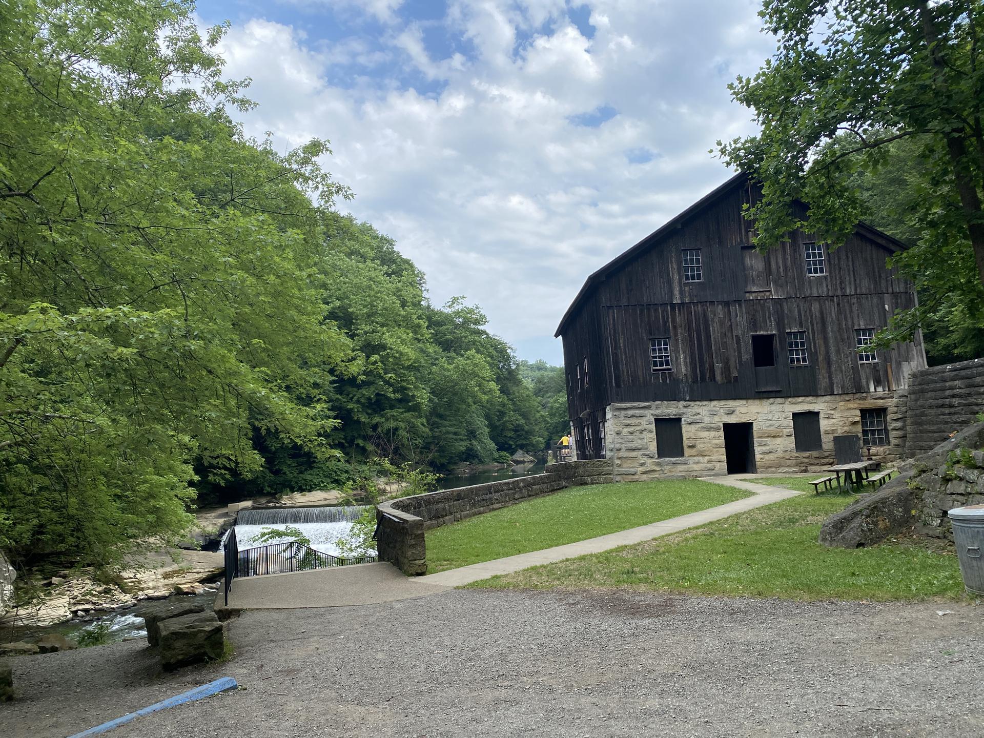An old wooden mill building stands beside a river with a small waterfall.